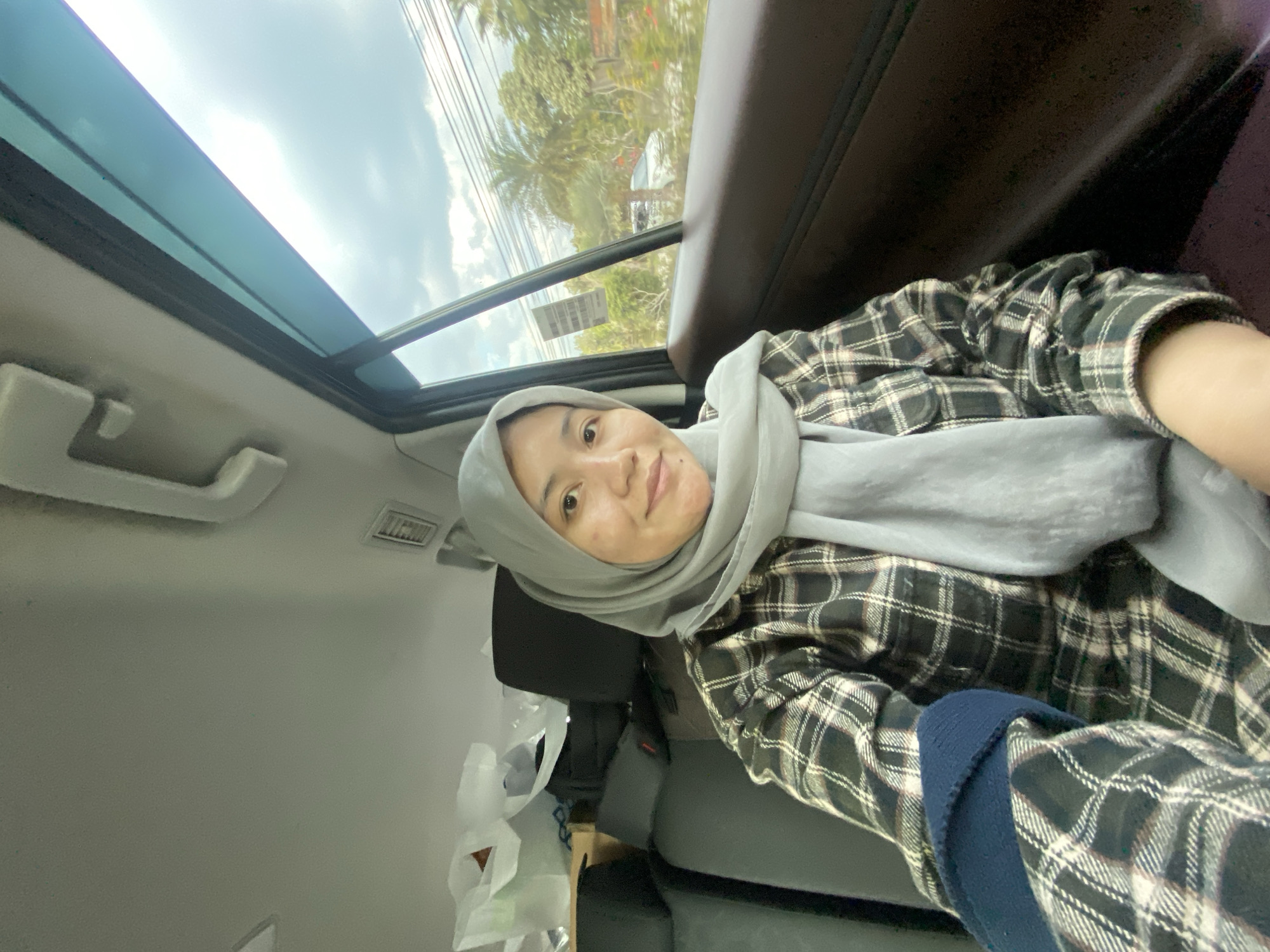 A medium-shot, self-taken photograph shows a woman in a car, looking at the camera with a slight smile. She is wearing a light gray hijab and a plaid shirt in shades of black, white, and reddish-brown. A dark blue sleeve is visible on her left arm. The interior of the car is visible, with dark gray seats and white bags and boxes in the background. Through the window on the right, trees and a white car are visible, suggesting an outdoor environment. The photo was taken on a bright, cloudy day. Based on visual cues such as the vegetation and common vehicle types, the image is likely from Banjarbaru, Indonesia.