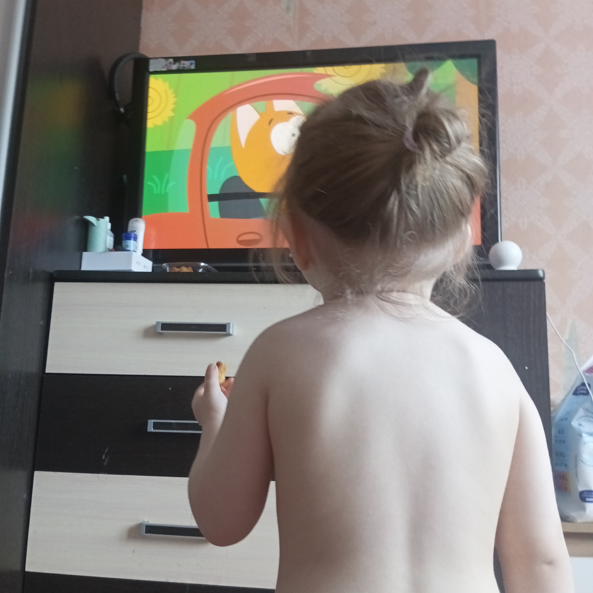 The scene depicts a child from behind, standing in a residential interior in Chaikovsky, Russia, focused on a television screen. The child's upper back and shoulders are bare, indicating a lack of a top garment, and their light brown hair is gathered in a bun secured with a pink hair tie. They hold a small, light brown, textured food item in their right hand.
The television, framed in black, displays a colorful animated program featuring a stylized orange, fox-like character with white ears and a large white eye, seated within an orange vehicle. The animated background is bright green with a yellow sun and small green plant details.
The television rests on a two-toned chest of drawers or media console. The top surface is dark brown. Below the television, the unit features two drawers: the top drawer has a light beige front and a dark brown frame, while the bottom visible drawer is entirely dark brown. Both drawers are equipped with horizontal silver handles.
Objects on the console surface include: a pair of black over-ear headphones hanging from the left side of the TV screen, a light blue container, and a white bottle with a blue cap on a white stand to the left of the screen. To the right of the screen, a small, white spherical object is present, along with a transparent container holding small, brownish snacks. A blue and white plastic package is partially visible on the far right. The wall behind the furniture is covered with light-colored wallpaper featuring a repeating diamond or grid pattern.