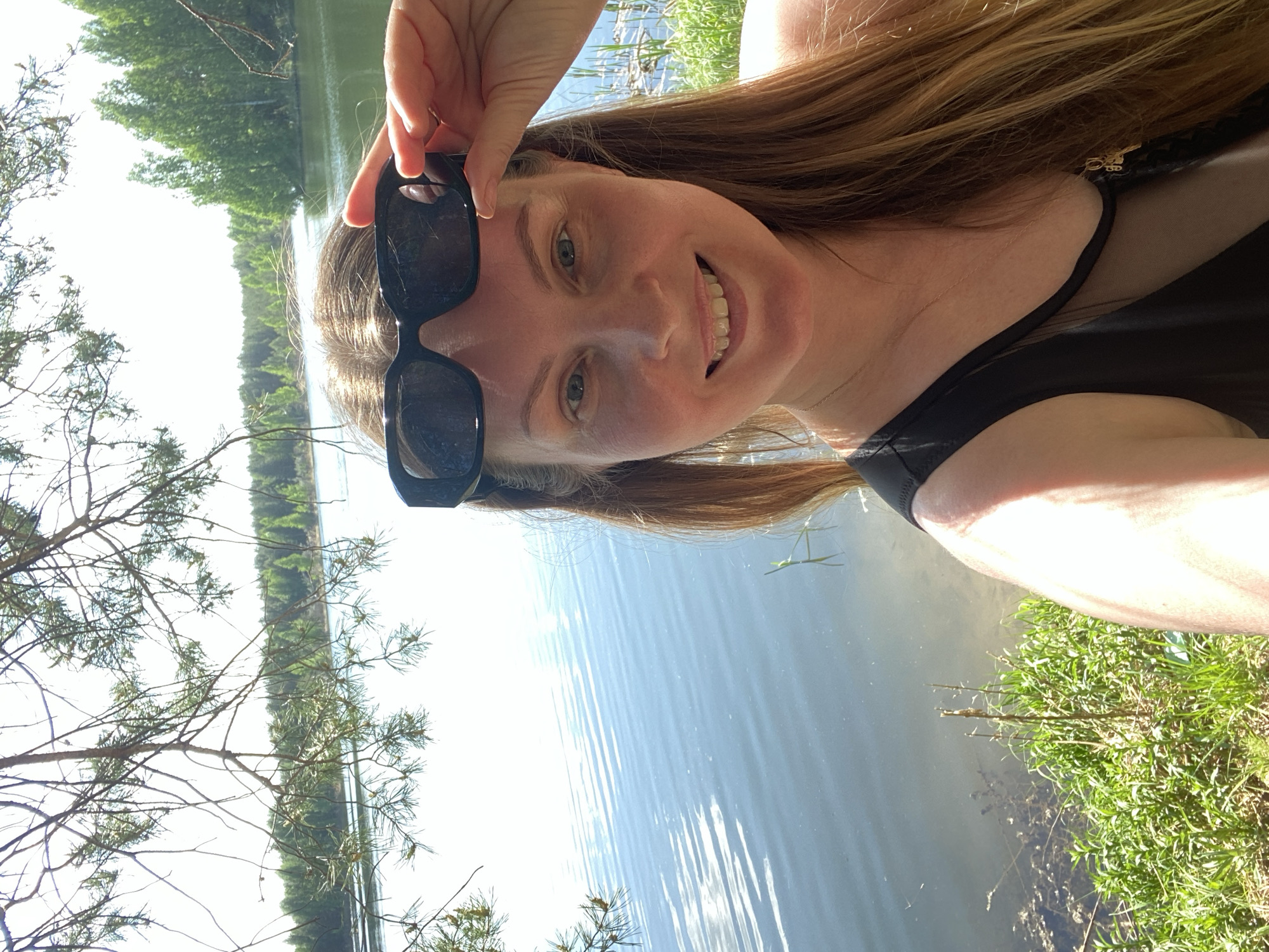 A woman stands by a body of water, possibly a lake or river. She is holding sunglasses above her eyes and smiling. The background includes trees and vegetation, typical of a natural environment. The image does not provide enough information to confirm the specific location in Novyi, Russia.