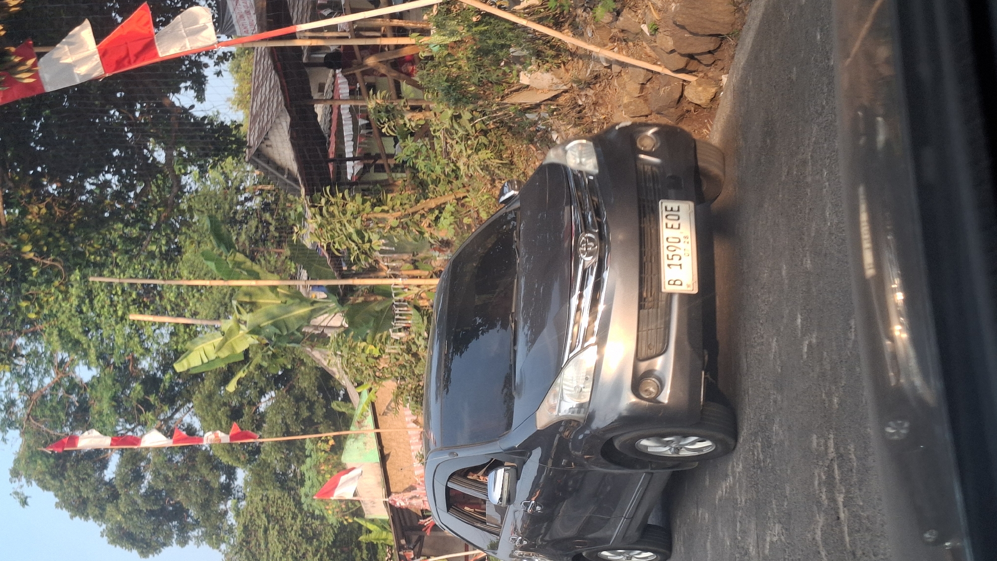 A dark gray sedan is parked on a paved road in Sirnagalih, Indonesia. The car's license plate reads "B 1590 EOE." The vehicle is angled slightly to the right, with its front bumper close to a rocky embankment. Behind the car, there are several small buildings with tiled roofs, lush green trees, and banana plants. Red and white Indonesian flags are visible on poles in the background. The sky is clear and bright, indicating daytime. The overall scene depicts a street view in a semi-rural or suburban area of Indonesia, possibly decorated for a national holiday.