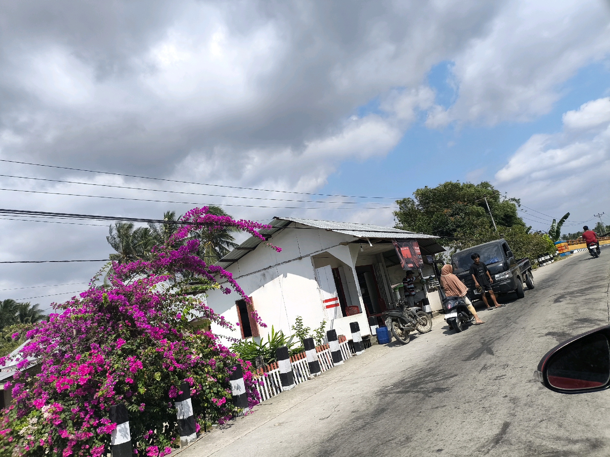 This is a street-level, eye-level, medium shot of a scene in Sido Mulyo, Indonesia, under a partly cloudy sky. The focal point is a white building with a corrugated metal roof, situated along a paved road. In front of the building, a vibrant pink bougainvillea bush is in full bloom, its branches cascading over a black and white striped fence.
To the right of the bougainvillea, the building has a covered porch area. A black pickup truck is parked in front of the building, and two people are standing near it. One person, wearing a light brown hijab and a beige top, is sitting on a motorcycle. Another person, a male wearing a black t-shirt and dark shorts, stands beside the truck. Several motorcycles are parked in the vicinity, including one in front of the bougainvillea bush and another next to the pickup truck. A blue barrel is visible near the motorcycle.
Further down the road, to the right, another person on a red motorcycle is riding away from the camera. In the background, there are green trees and more palm trees. Power lines are strung across the top of the frame. A red wing mirror, likely from the vehicle the photo is taken from, is visible in the bottom right corner. The road surface shows some dark stains and tire marks.