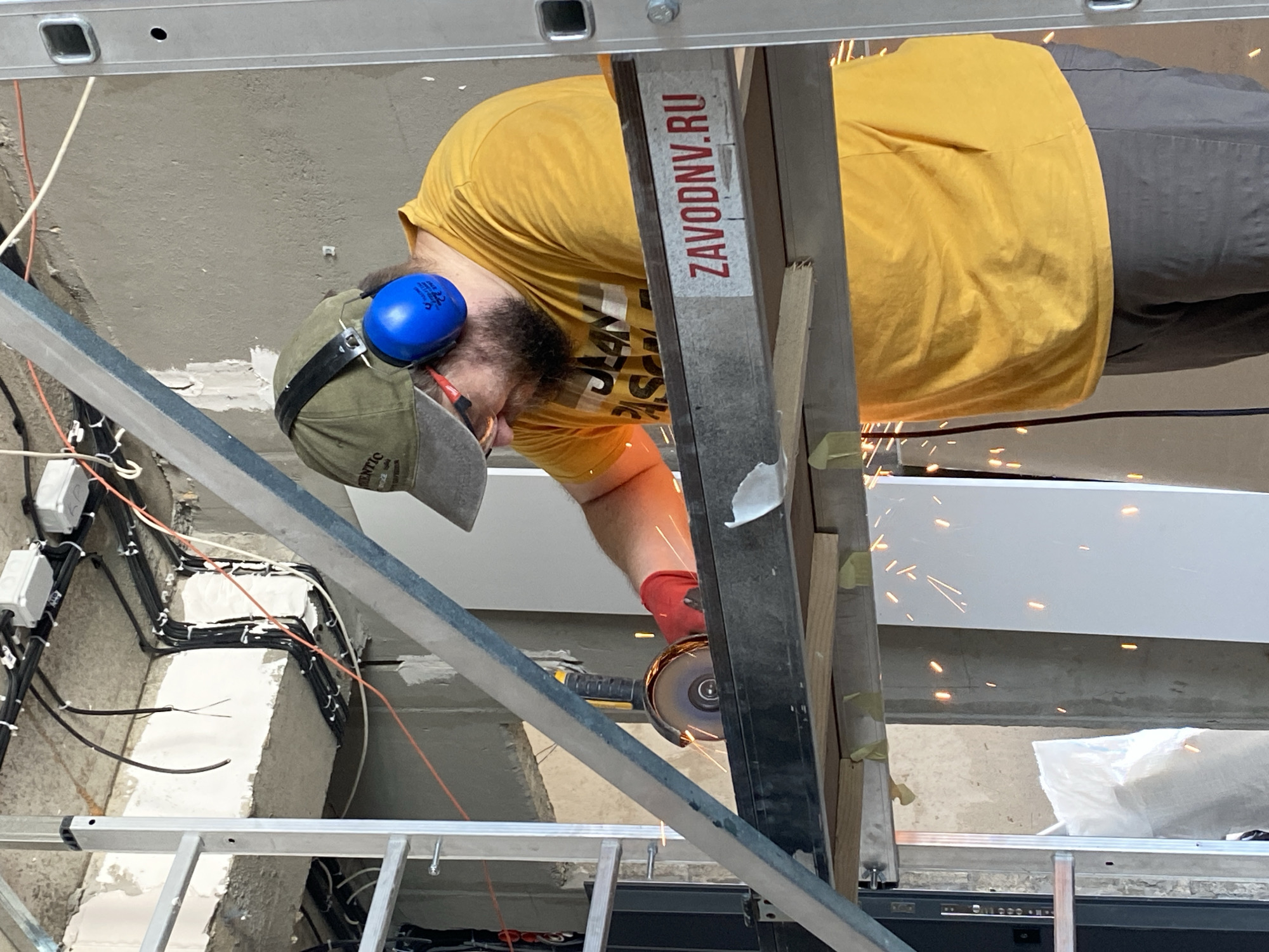 The image shows a construction worker wearing a yellow t-shirt, grey pants, a green cap, red gloves, and blue ear protectors. He is using an angle grinder to cut a metal beam, which is producing sparks. The worker is standing on a temporary metal platform. The background reveals exposed concrete walls, electrical wiring, and scaffolding, indicating an ongoing construction or renovation project. The scene suggests an indoor construction site, possibly in Batumi, Georgia, given the context of previous images.
