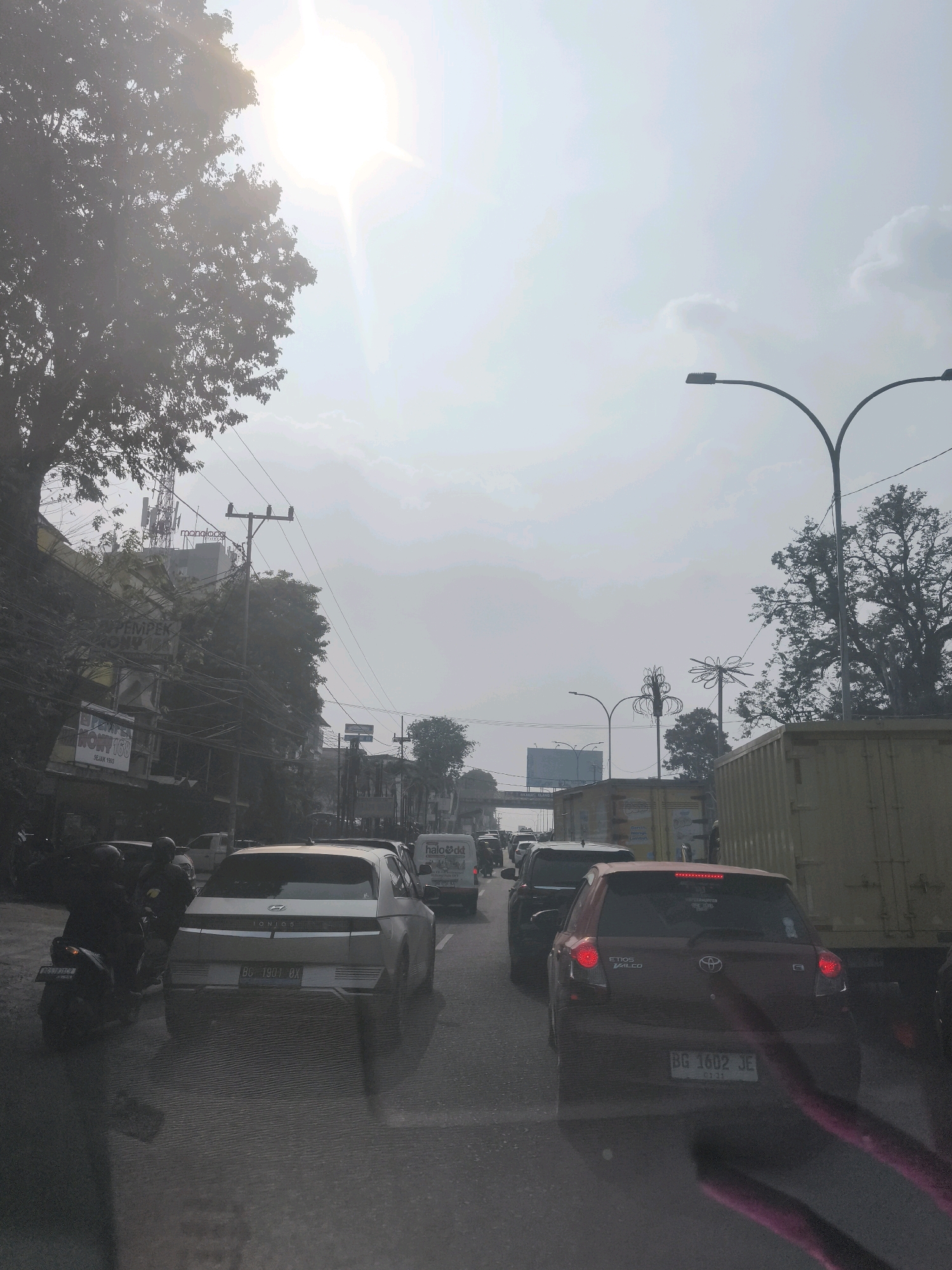 The image depicts a congested traffic scene in Palembang, Indonesia, likely on a daytime with hazy sky and bright sunlight filtering through. The foreground is dominated by a road with several vehicles, including a white Hyundai Ioniq and a red Toyota Etios Valco. A motorcycle with two riders is also visible on the left. Behind the cars, a large yellow shipping container truck is parked. In the background, trees and buildings line the street, with power lines and streetlights also present. A prominent sign with the text "monologes" is visible on a building. The overall atmosphere suggests a typical urban traffic situation in Palembang.