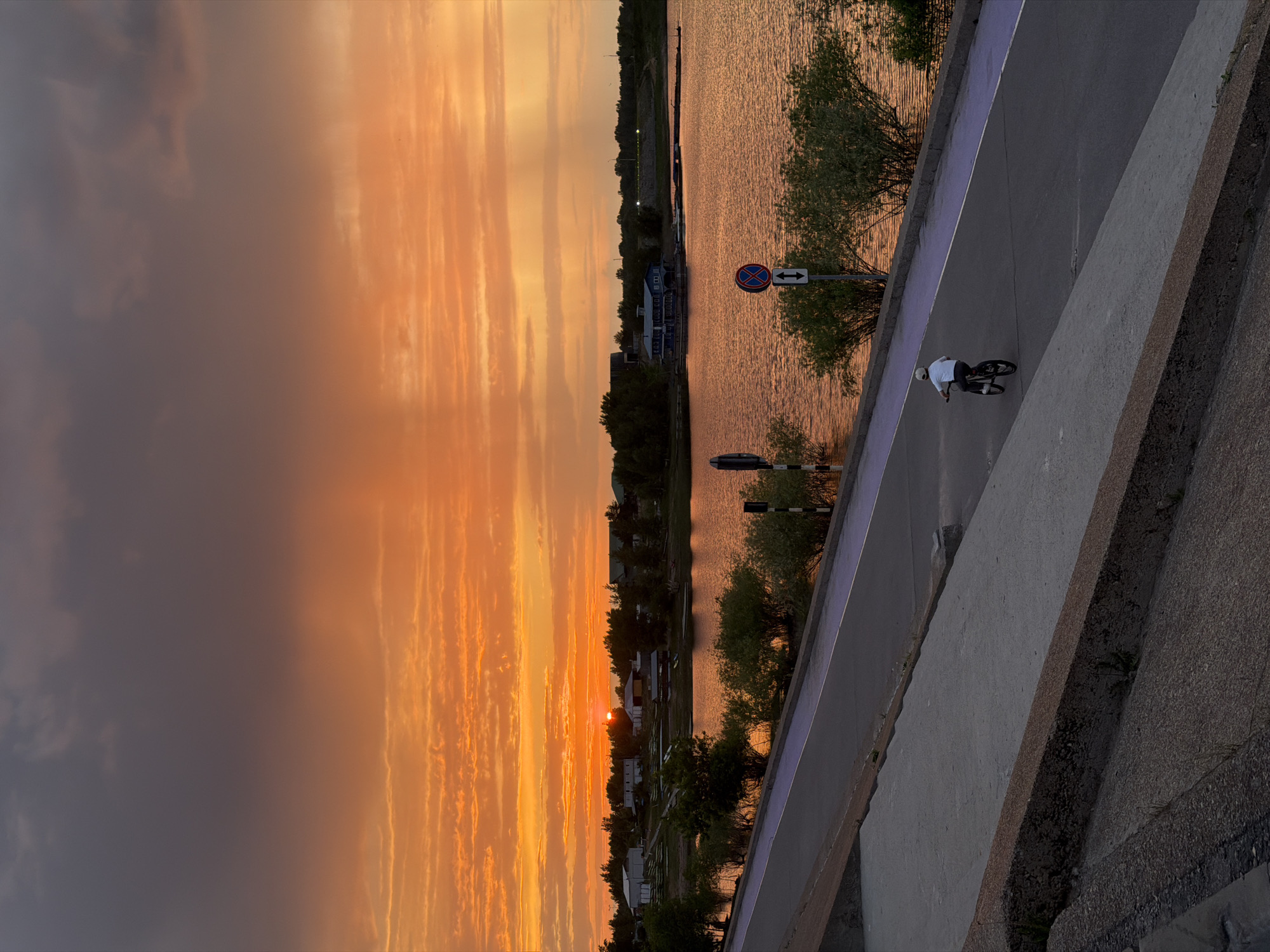 The image shows a sunset scene near a body of water, possibly the Volga River, in Nizhny Novgorod, Russia. The sky displays a vivid orange gradient, indicative of either sunrise or sunset, with cloud formations streaking across the frame. The river reflects the warm light. A cyclist is riding along a path beside the river, along a concrete barrier. Signage and trees line the riverbank, and buildings appear in the distance.