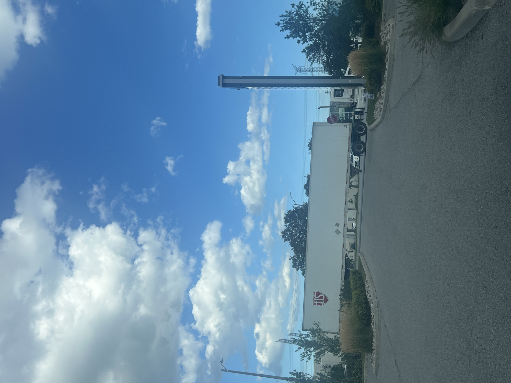 In Vaughan, Canada, under a bright blue sky with scattered white clouds, a large white semi-trailer truck with "TIS" logo on its side is parked. The truck is positioned in a paved area, possibly a parking lot or industrial zone, with a road leading away from it. To the right of the truck, a tall, slender, modern gray and white sign structure stands, displaying some text. A red stop sign is visible on a pole near the truck's cab. Trees and green foliage surround the area, with a large, tall transmission tower visible in the distance. The overall scene suggests an industrial or commercial setting.