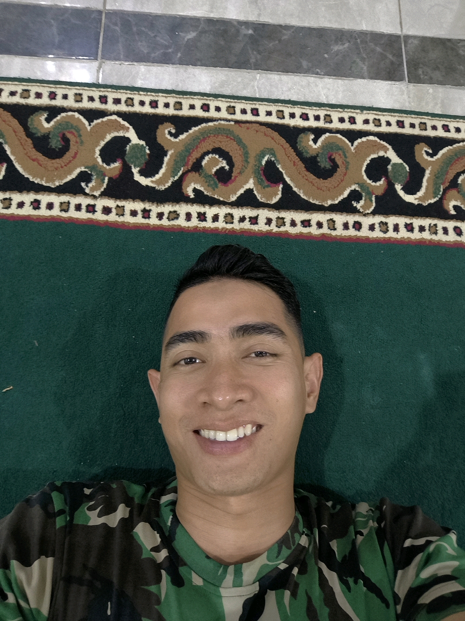 A man in a camouflage t-shirt smiles at the camera, lying on a green carpet that borders a patterned rug. The rug is adjacent to a tiled floor. The man's hair is short and dark, and he has a light complexion. The overall lighting suggests an indoor environment.