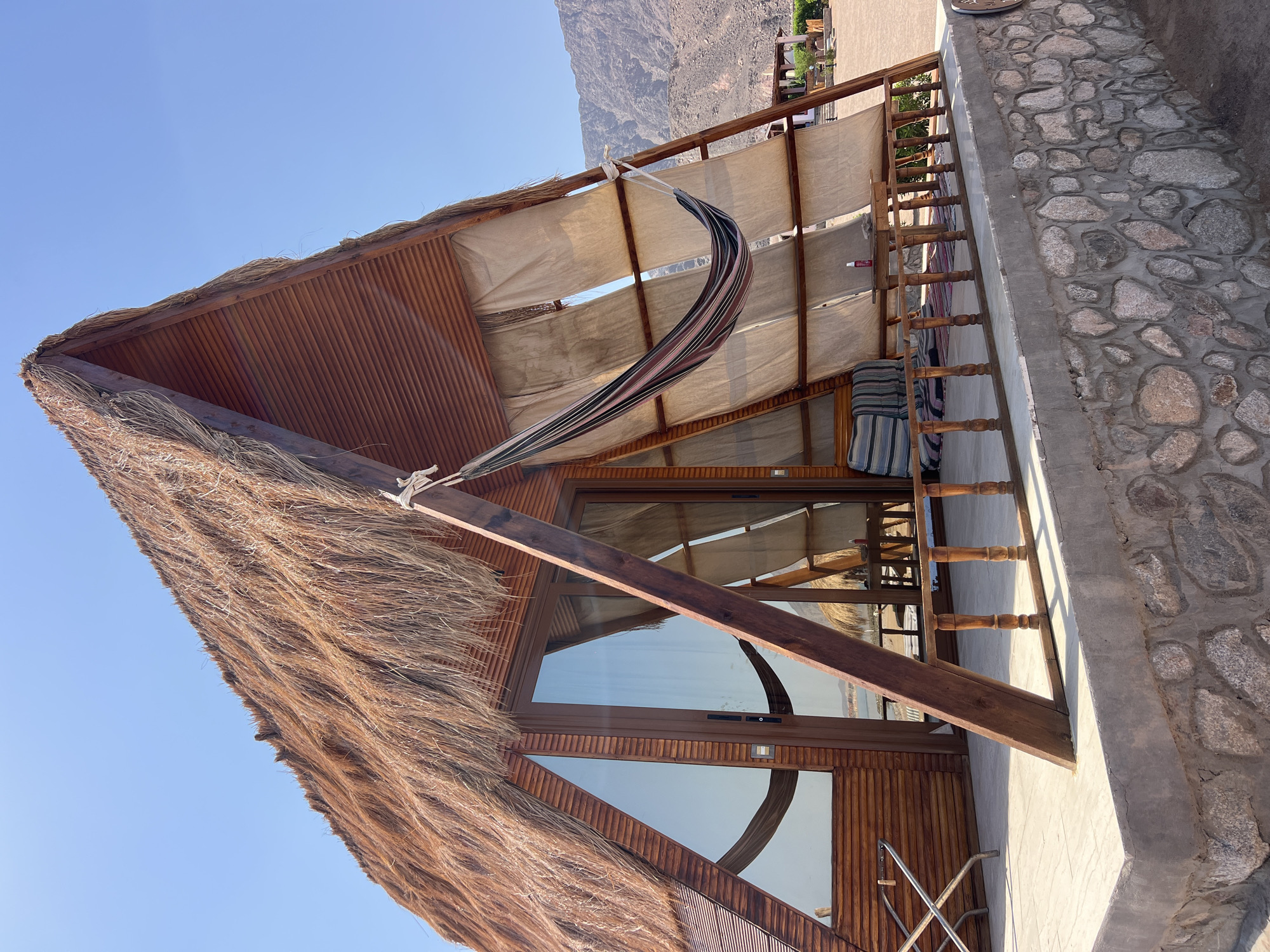 The image shows a triangular wooden cabin with a thatched roof, situated in a sunny outdoor environment. The cabin has large glass doors and windows, reflecting the clear blue sky and a distant body of water. A hammock is suspended on the covered porch area, which is furnished with a few striped cushions and a small wooden table. The cabin is built on a stone foundation and overlooks a sandy area with some scattered vegetation and distant mountains. The overall scene suggests a resort or glamping location in a warm climate.