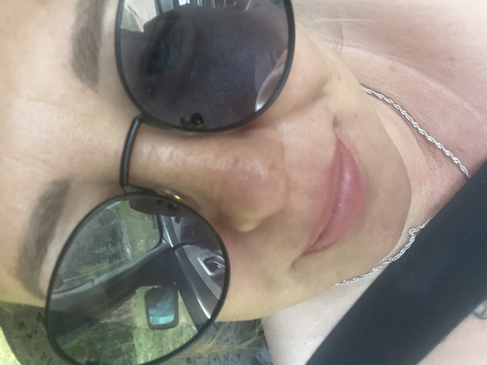 An adult Caucasian female is depicted from the neck up, turned toward her left, wearing sunglasses. The sunglasses have a dark tint, reflecting a car interior and roadside foliage. She also wears a fine, silver-colored necklace. The location cannot be determined as Chaikovsky, Russia, based solely on the provided image.