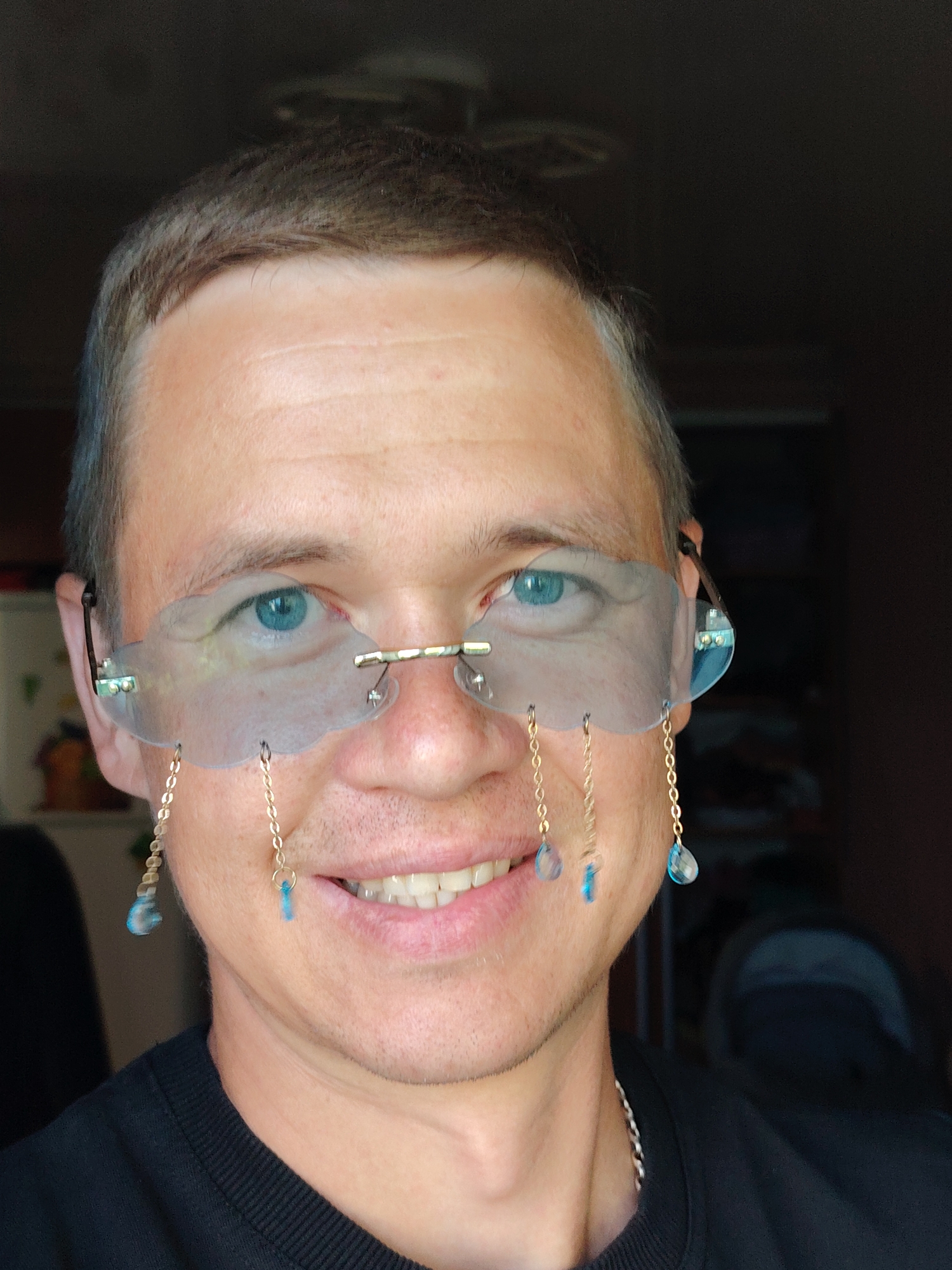 A medium shot captures a Caucasian man with short, dark brown hair and blue eyes, wearing a black t-shirt. He is wearing unusual, rimless glasses shaped like clouds, with gold chains and blue droplet-shaped charms hanging from them. The man is smiling, revealing his teeth. The background is dark and out of focus, suggesting an indoor setting. The image was taken in Chaikovsky, Russia.