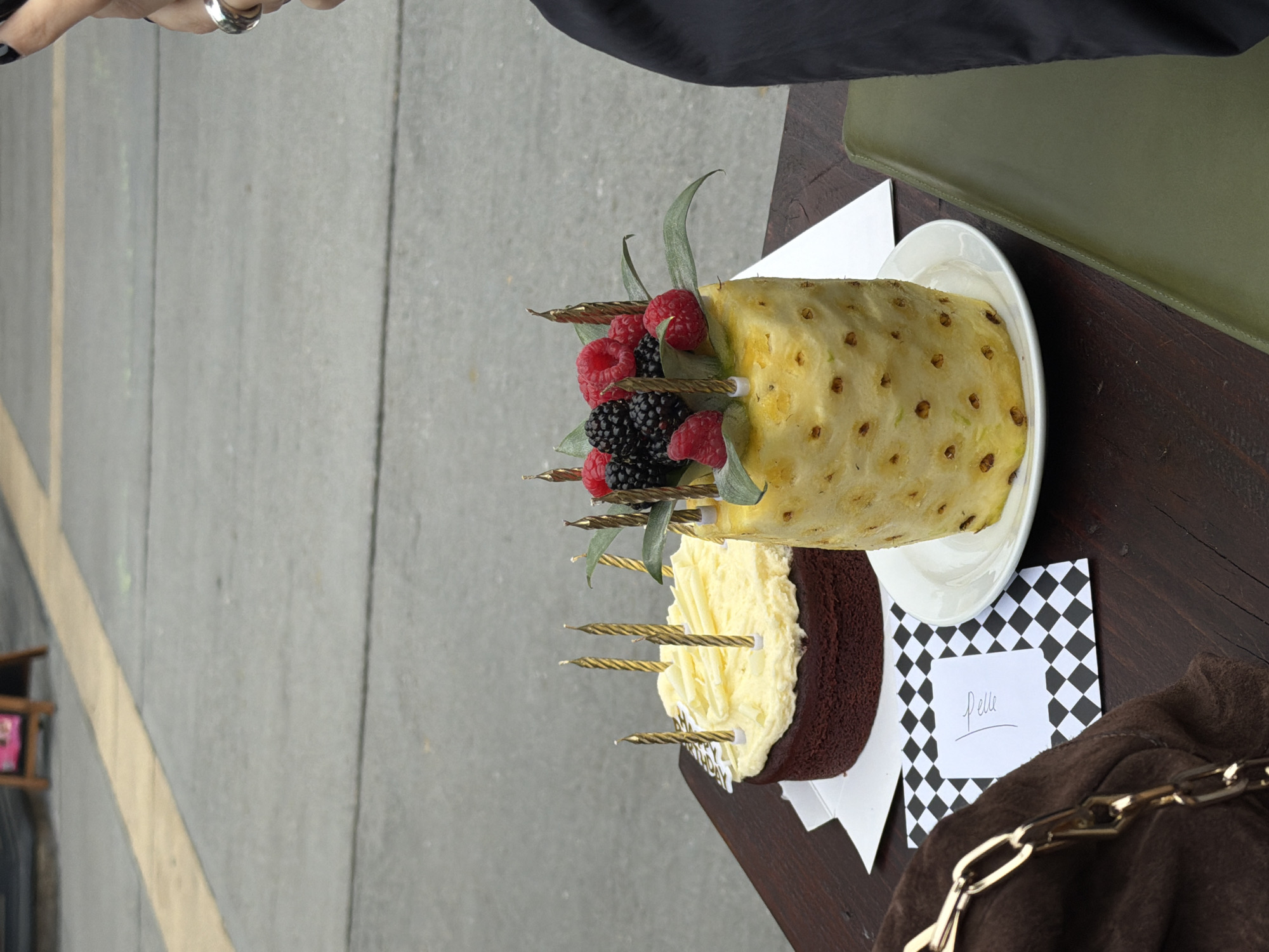 The image shows a birthday cake, partially made of pineapple, topped with raspberries and blackberries, with golden candles inserted. The cake sits on a white plate and is accompanied by a small card with the name "Pelle" written on it, placed on a black and white checkered background. The setting is outdoors, on a dark wooden table next to a street or sidewalk. A portion of the arm and hand of a person with rings is visible in the upper left of the frame. Part of a dark jacket or coat can also be seen. Based on the attire and style of the setting, and the general aesthetic, it is plausible that the image was taken in the city of London, United Kingdom.