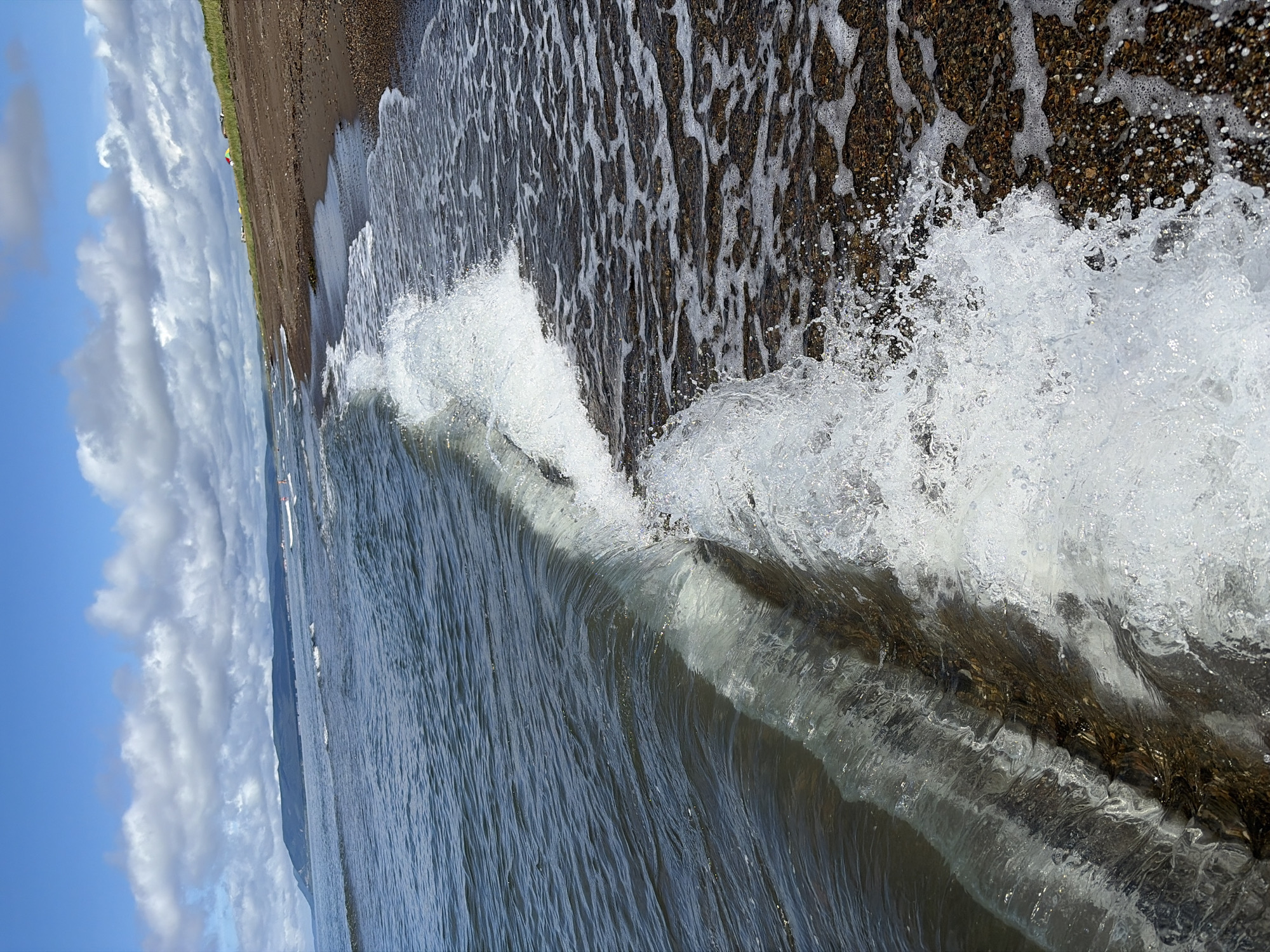 The image depicts a coastal scene with a wave breaking on a pebbly beach under a partly cloudy blue sky. The water is a deep blue-green, with gentle ripples on the surface. The wave is captured mid-crest, with foam and spray arcing forward. The beach is composed of dark pebbles and sand, with some seaweed present. In the distance, a low, green-covered hill or mountain range forms the horizon line. A few scattered buildings or tents are visible along the coastline in the far distance, and a white boat is present on the water. The overall impression is one of a natural, serene coastal environment. The location in Ozerskoe, Russia, is noted as a coastal area.