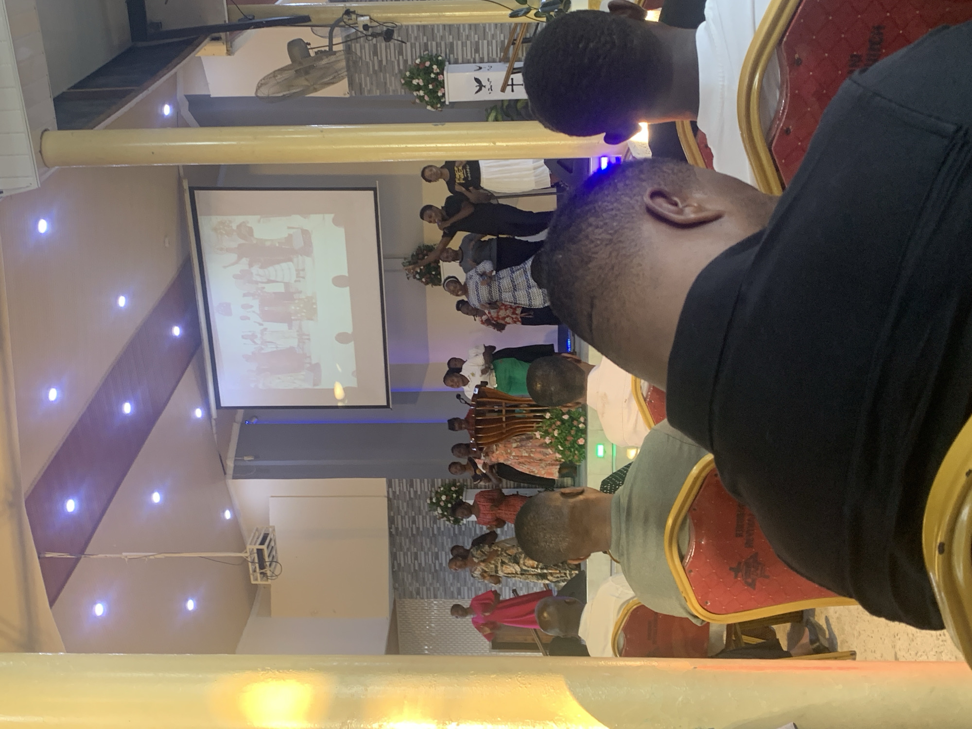 A group of people are gathered on a stage in a large indoor space, possibly a church or auditorium, as observed from the audience. A projector screen displays an image behind them. The event appears to be taking place in Dar es-Salaam, Tanzania, based on the visual information available. Attendees are seated in red and gold chairs.