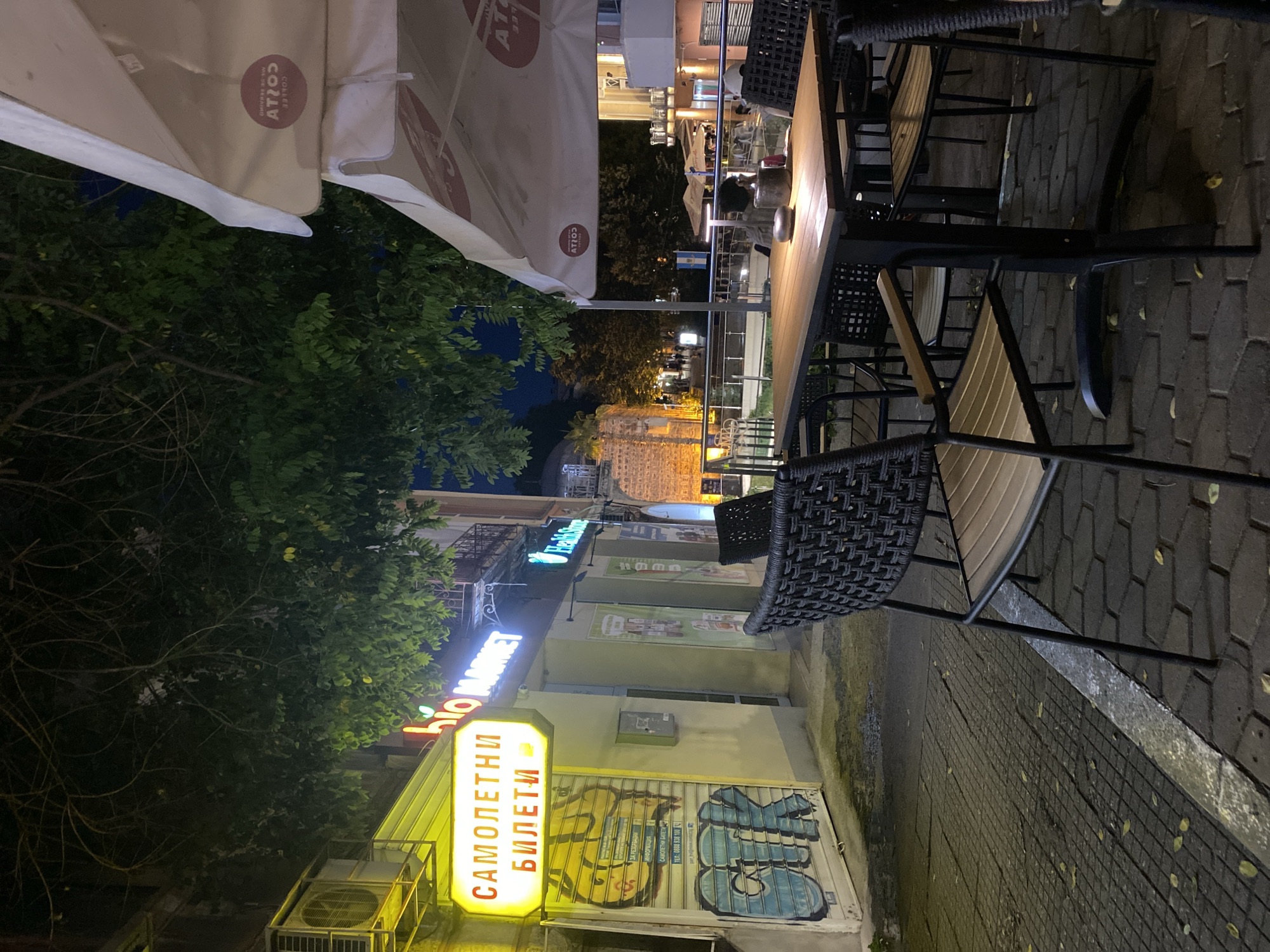The image depicts a street-level view of an outdoor seating area for a cafe or restaurant in Plovdiv, Bulgaria, at night. In the foreground, a wooden table is set with chairs featuring dark woven backs and beige seats. A small lamp illuminates the table, on which rests a metallic object, likely an ashtray, and a bottle. Beyond the immediate seating area, other tables and chairs are visible, some occupied by patrons.
To the left, a building facade is adorned with a brightly lit yellow sign reading "САМОЛЕТНИ БИЛЕТИ" (Airplane Tickets) in Cyrillic. Below this sign is a roll-up metal door with graffiti, including large blue and yellow letters forming the word "CRK." Adjacent to the ticket shop, a store with a neon blue sign reading "bio MARKET" is visible, with further storefronts and signs extending into the background.
Overhead, dense foliage from trees partially obscures the night sky and the upper portions of the buildings. Umbrellas, possibly belonging to the cafe, are visible in the upper right portion of the image, one bearing a circular logo with text that appears to read "COSTA COFFEE." The ground is paved with interlocking bricks and cobblestones, with scattered fallen leaves. The overall ambiance suggests a lively evening setting in a commercial district of Plovdiv.