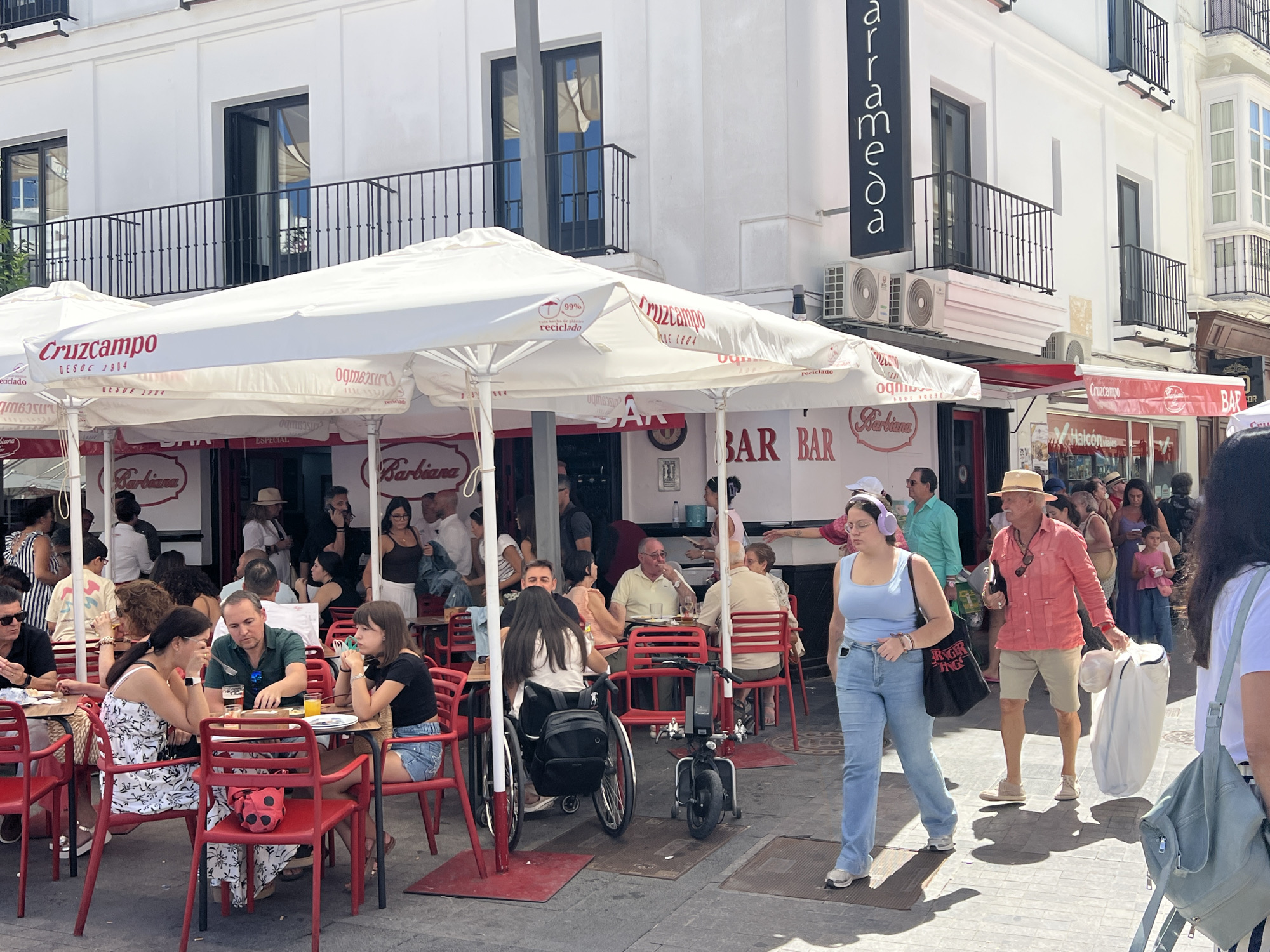 The image depicts an outdoor dining scene at a cafe or restaurant in Sanlúcar De Barrameda, Spain. Multiple people are seated at tables under large white umbrellas with "Cruzcampo" branding. The architecture suggests a Spanish setting with white buildings and balconies. In the foreground, a woman in a blue tank top and jeans walks past, wearing headphones. To her right, an older man in a coral shirt and beige shorts is walking, carrying a white bag. A woman in a wheelchair is visible at one of the tables. The overall atmosphere is that of a busy urban street with people enjoying a meal and drinks outdoors.