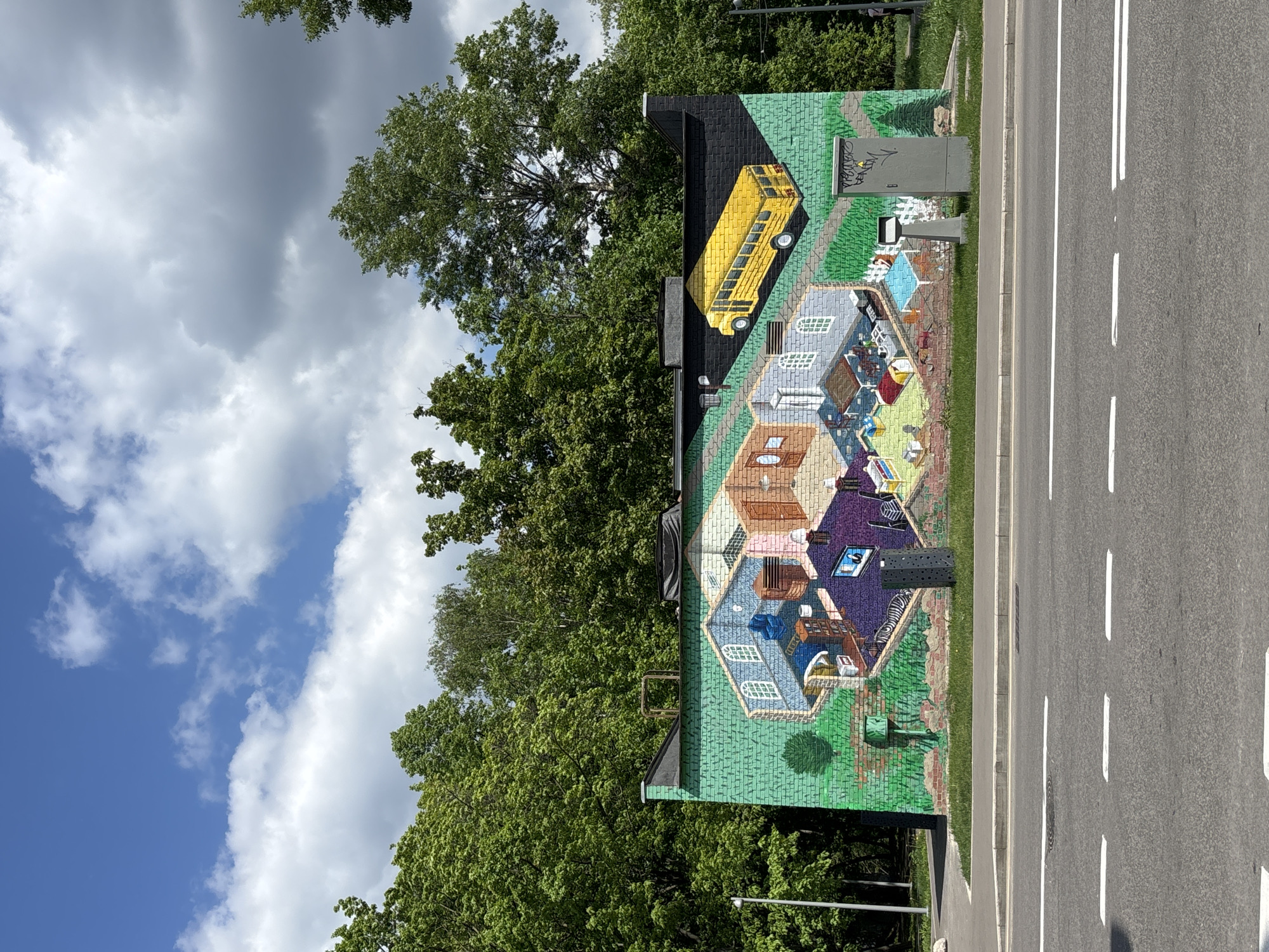 The image depicts a large mural painted on the side of a building. The mural features an isometric-style rendering of interconnected rooms and a yellow school bus on the roof. A road with white lane markings is visible on the right side of the frame. Trees and a cloudy sky form the backdrop. The building appears to be located in an urban environment, possibly Moscow, Russia, given that it is the prompt's subject matter. The mural is next to an electrical box with what seems to be some graffiti on it.