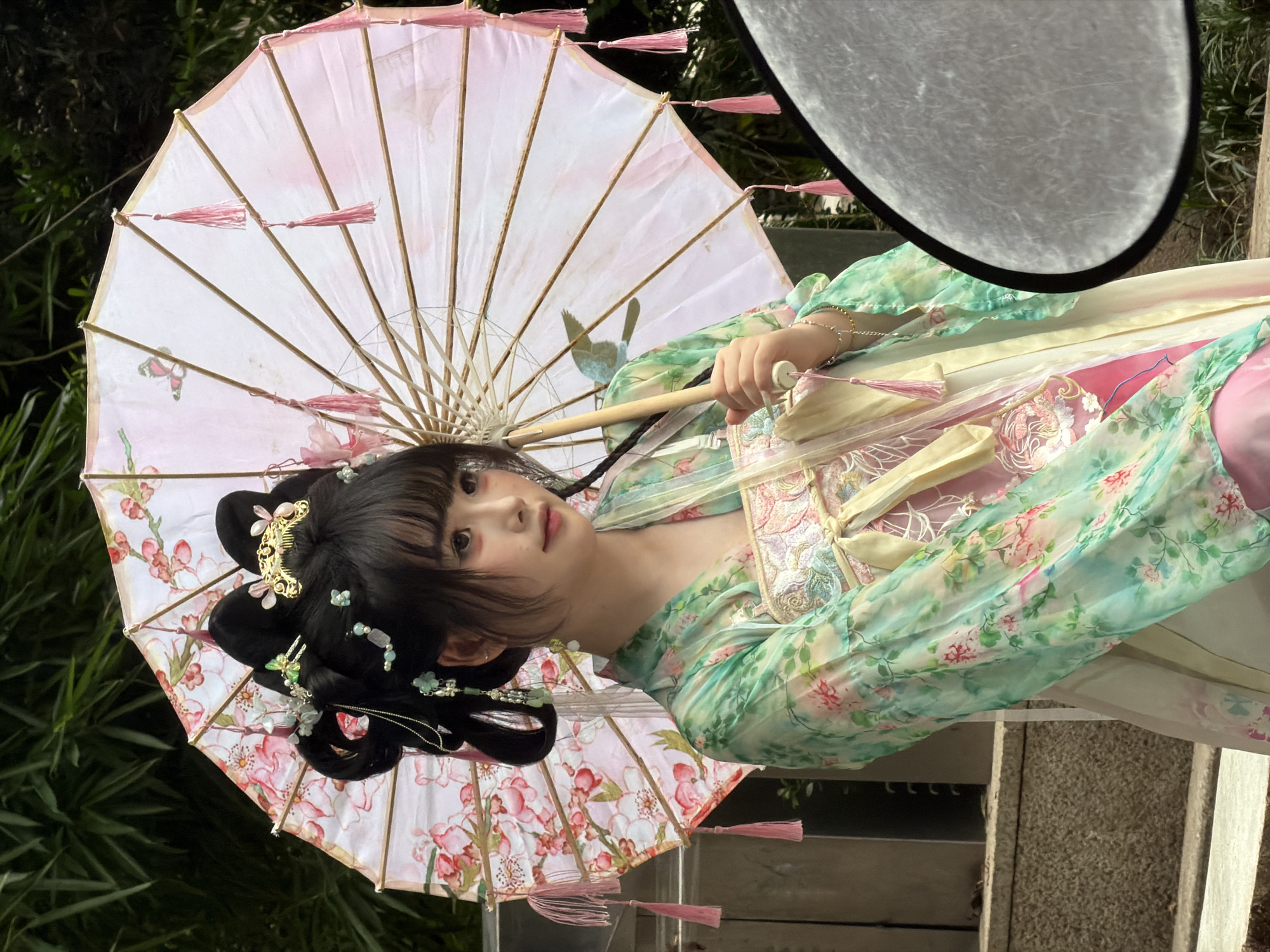 A young woman in traditional Chinese attire stands outdoors, holding a decorative umbrella. The umbrella is adorned with floral patterns and pink tassels. Her outfit consists of a light green, flowing robe with intricate embroidery and a pink undergarment. Her hair is styled in an elaborate updo with hairpins and decorative accessories. The background features lush greenery, suggesting a garden or park setting. The woman's makeup is subtle, with a focus on her eyes and cheeks. A reflector disc is partially visible in the lower right corner of the frame, indicating a photoshoot is in progress. The scene appears to be located in Yuzhong District, China, known for its historical and cultural sites.