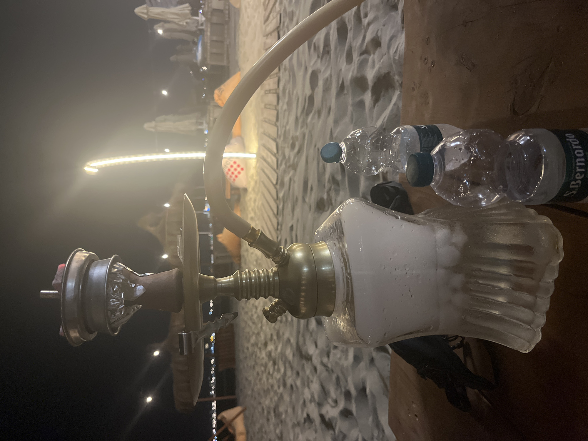 This media file captures a night scene at a beachside establishment, likely a cafe or lounge. The primary focus is a hookah pipe, made of brass and glass, filled with what appears to be a white, smoky substance. The glass base of the hookah is filled with water and ice, creating condensation on the outside. A light-colored hose extends from the hookah, disappearing out of frame.
Two plastic water bottles are visible on a wooden surface next to the hookah. One bottle has a dark green cap and is labeled "S. BERNARDO", suggesting it is San Bernardo water. The other bottle has a blue cap. The surface appears to be a table or ledge.
In the background, the sandy beach is visible, with the gentle slope of the sand suggesting a shoreline. Several umbrellas, likely for beach use, are present, some illuminated by artificial lights. String lights are also visible, casting a warm glow and creating bokeh effects. The overall atmosphere is dim, indicating it is nighttime. The weather appears to be calm, with no strong winds suggested by the scene.
The setting evokes a relaxed, social environment, possibly for evening leisure. The presence of the hookah and water bottles suggests a gathering or a moment of personal relaxation. The overall mood is tranquil and ambient.
The location context places this scene in Durrës, Albania, a coastal city known for its beaches and nightlife. This information aligns with the visual cues of a beach setting at night.