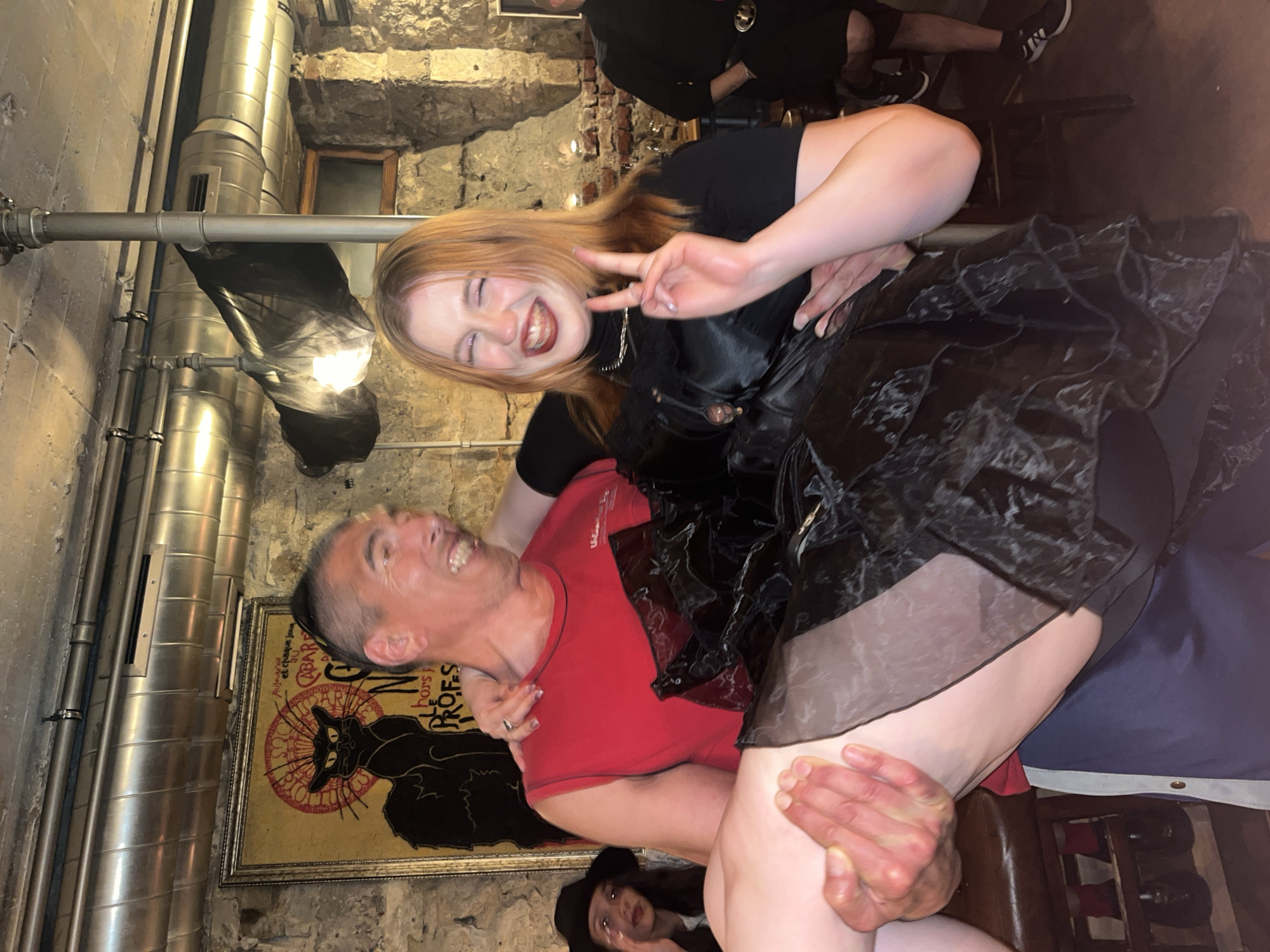 A man in a red shirt is holding a woman with red hair, wearing a black top and black skirt, in his arms. The woman is giving a peace sign and smiling. They are inside what appears to be a pub or bar with stone walls and exposed pipes. There's a framed poster with a black cat on the wall behind them. People are visible in the background. The location is Varna, Bulgaria.