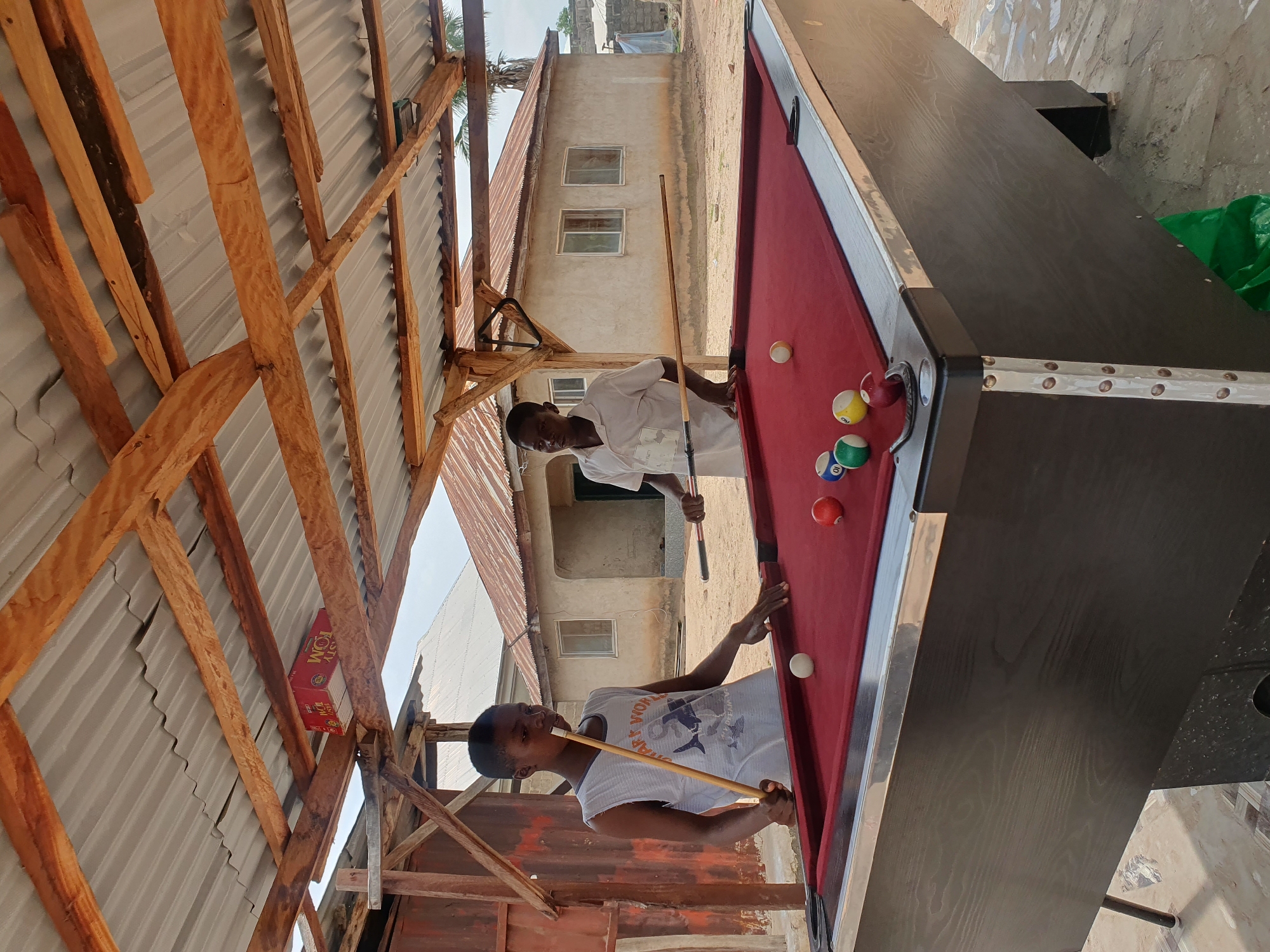 Two young males are playing a game of billiards outdoors in Lokoja, Nigeria, under a makeshift shelter. The shelter's roof is made of corrugated metal sheets supported by rough-hewn wooden beams. The ground is paved with irregular stones, and the background shows a sparsely built environment with modest residential buildings and a palm tree.
The male on the left, wearing a sleeveless white t-shirt with a graphic print, is positioned on the left side of the billiard table, holding a cue stick and aiming at a white ball. His body is angled towards the table, indicating active participation in the game. The male on the right, dressed in a white short-sleeved shirt, stands on the right side of the table, also holding a cue stick and looking towards the table.
The billiard table is covered with a red felt surface. Several billiard balls are visible on the table, including a white cue ball, a solid red ball, a striped ball numbered 10, a striped ball numbered 6, and a striped ball numbered 9. The table itself has a dark wooden exterior and metal trim around the edges.
In the background, a light-colored residential building with a corrugated metal roof and visible windows stands under a cloudy sky. The overall scene suggests a casual recreational activity taking place in a residential or community area within Lokoja.