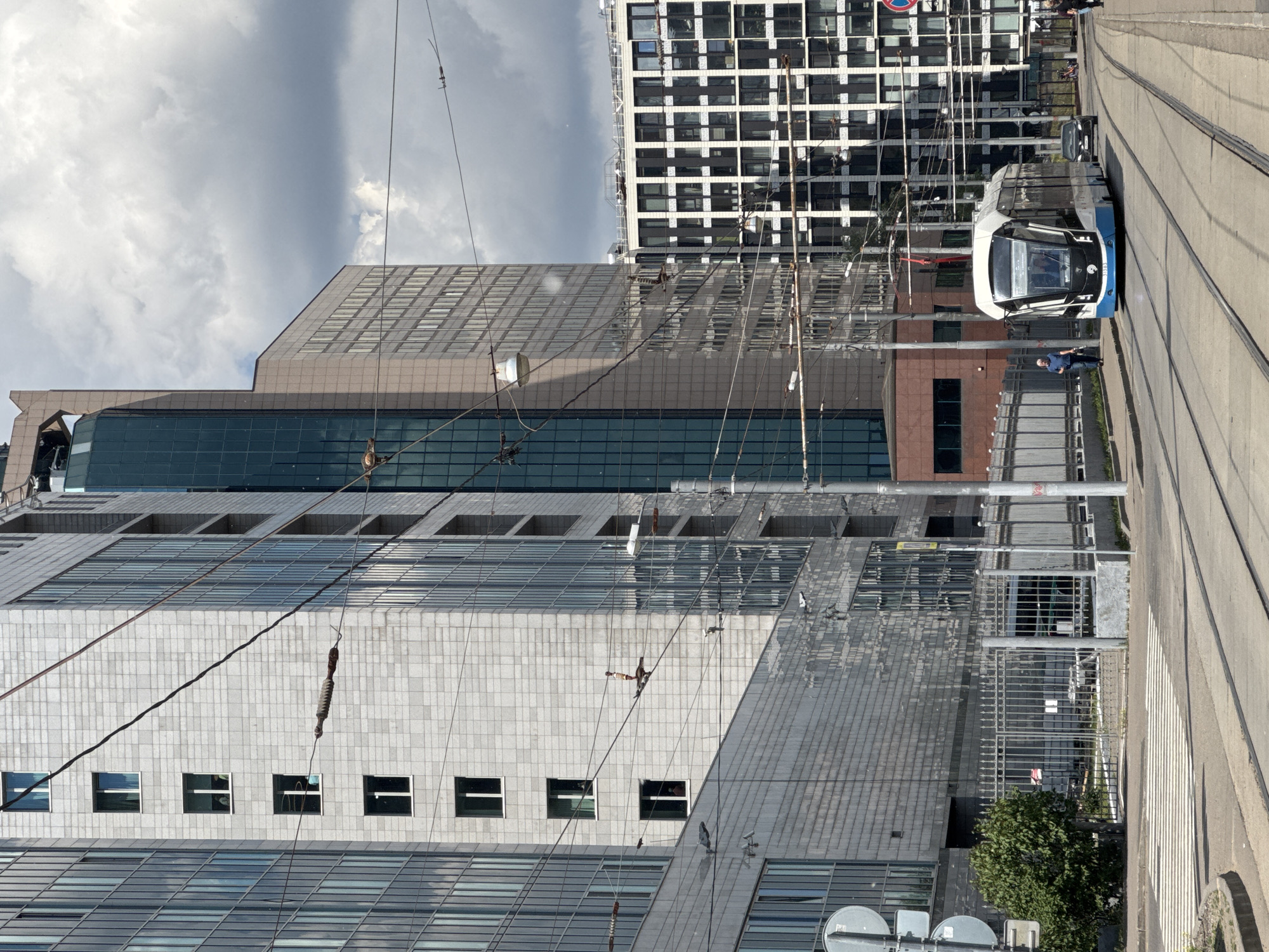 This image captures an urban scene in Moscow, Russia, during the daytime under a cloudy sky. The main subject is a modern, white and blue tram situated on tracks, either stopped or moving slowly, towards the right side of the frame. A network of overhead electrical lines, essential for the tram, traverses the sky above the street.
Parallel to the tram tracks, a sidewalk is visible where a man in a blue shirt stands near the tram. Other pedestrians can be seen further down the sidewalk.
The background is dominated by several large, contemporary buildings. One prominent building on the left features a light grey facade with numerous square windows and large reflective glass panels on its lower and upper sections. Another building behind it has a distinctive black and white checkered pattern created by its windows and structure. These modern buildings reflect the cloudy sky and surrounding elements.
A metal fence runs alongside the street, and a small green tree provides a touch of nature amidst the urban landscape. The scene conveys a typical city environment with modern infrastructure and public transport. The weather appears overcast but bright, with significant cloud cover. No discernible text is visible on any signs or buildings.