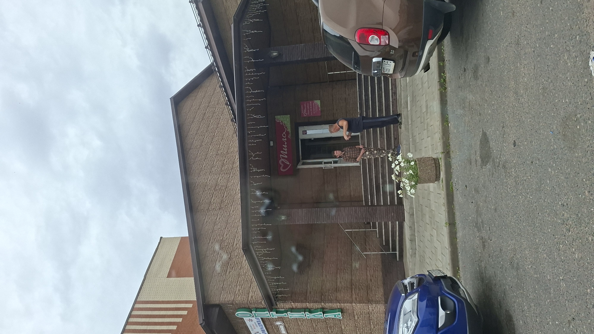 The image captures a street-level view of a building's entrance in Myadzyel, Belarus. The building features a brown brick facade with a covered entryway adorned with string lights. To the left of the entrance, a vertical green sign reads "ОПТИКА" (Optika), indicating an optical shop. Directly above the entrance, a pink sign with a heart logo and the word "Мила" (Mila) is visible, likely a cosmetic or beauty store.
Two individuals are standing on the steps leading into the building. The person on the left appears to be a younger male wearing a plaid shirt and camouflage pants. The person on the right is an older male with a sleeveless shirt and dark pants, gesturing with his hands as if in conversation.
In the foreground, a blue car is parked to the left, and the rear of a brown car is parked to the right. A potted plant filled with white flowers sits on the sidewalk in front of the steps. The ground is paved with light-colored rectangular stones, and the street in front is asphalt. The sky is overcast with gray clouds.