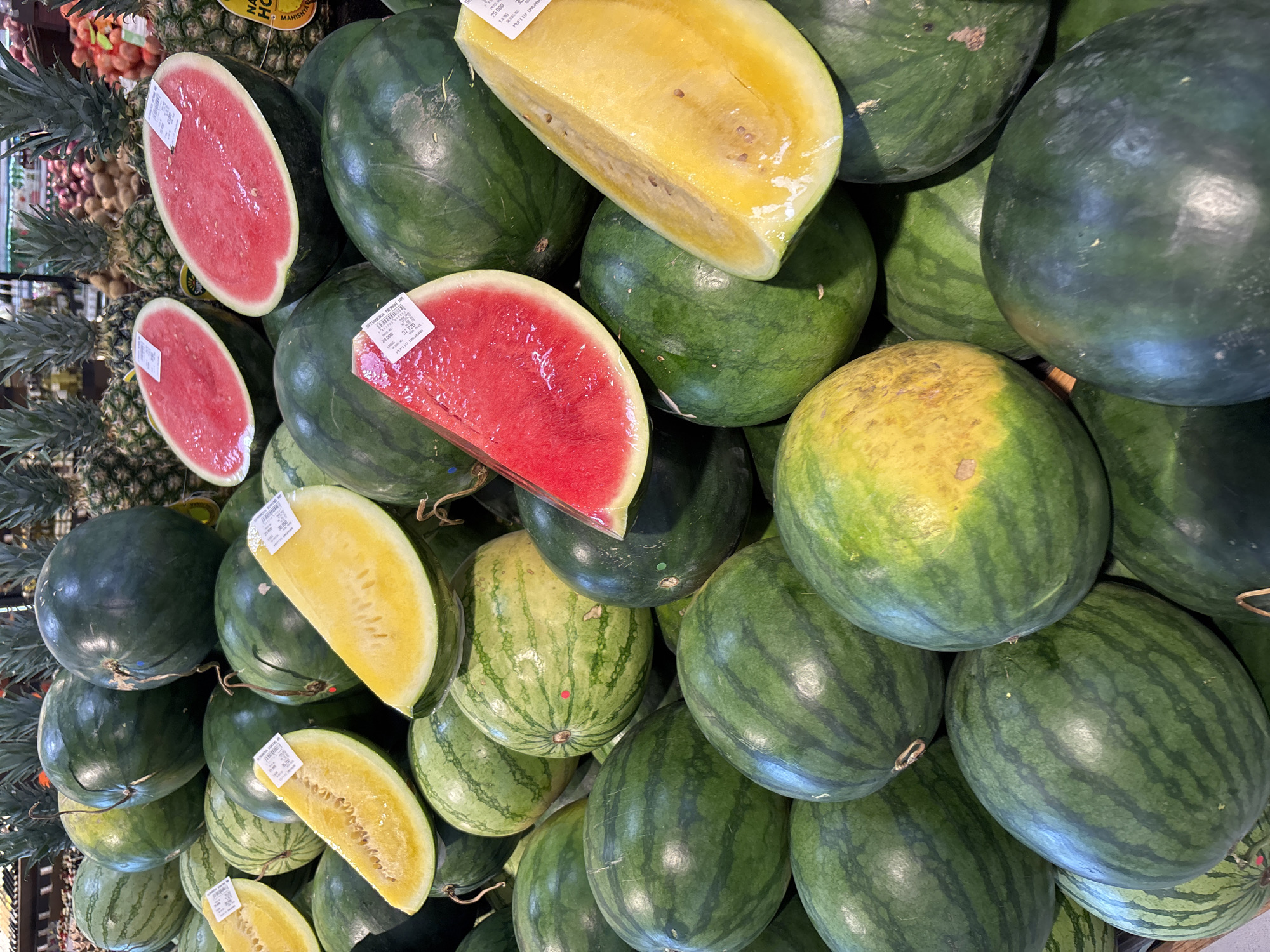 A high-angle shot depicts a heap of watermelons, both whole and cut, at a market stand in Kuta Selatan, Indonesia. The uncut watermelons vary in color from dark green to green with light yellow patches and stripes. Several cut watermelon halves, wrapped in clear plastic, display red and yellow flesh varieties. Price tag stickers are visible on a few of the watermelons. Pineapples are visible in the background.