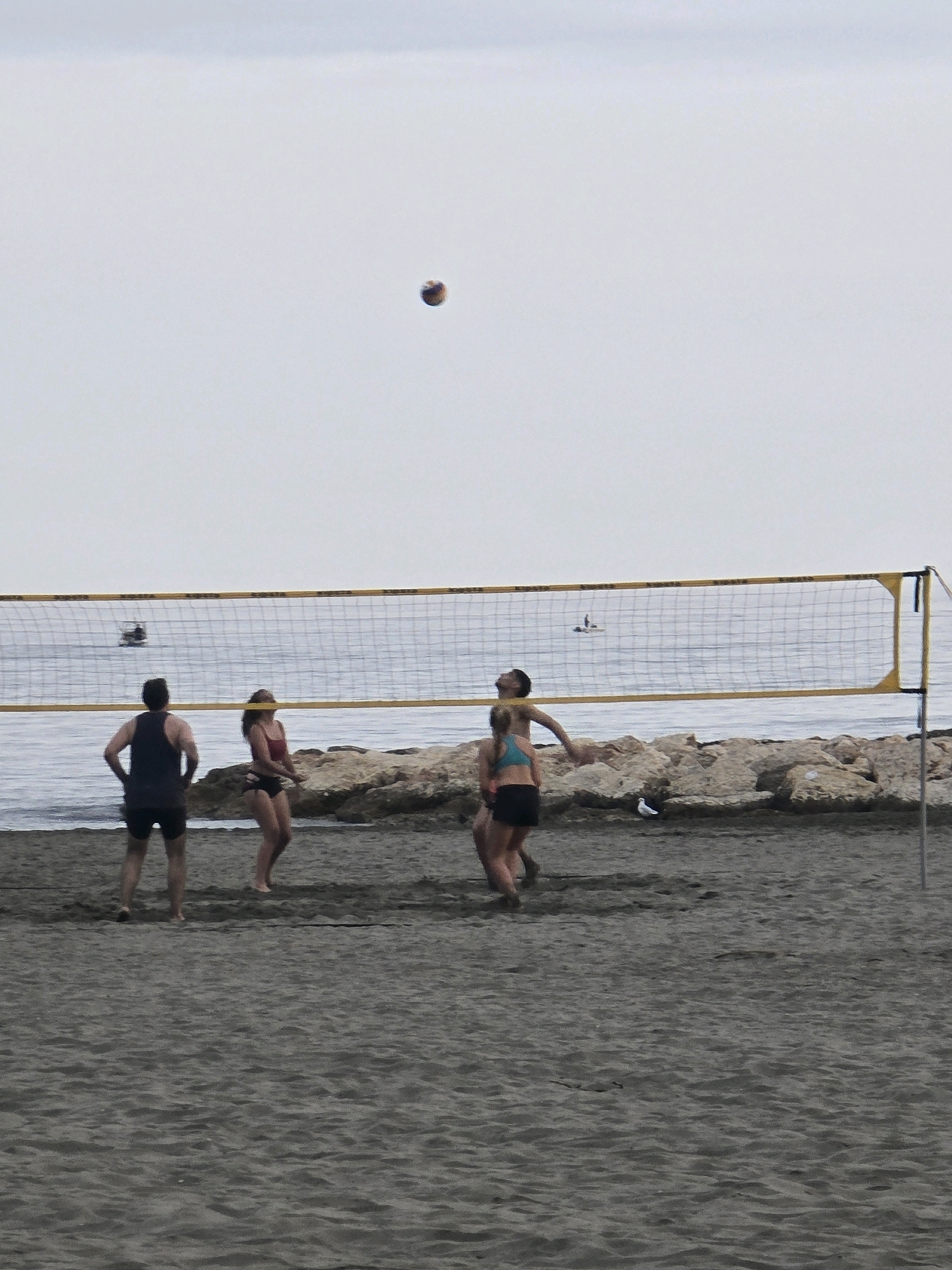 The image depicts a beach volleyball game on a sandy beach, likely in Málaga, Spain, given the presence of the Mediterranean Sea and what appears to be a rocky coastline. The sky is overcast. A yellow-trimmed volleyball net stretches across the sand.
Four individuals are actively playing:
- On the left, a man in a dark tank top and black shorts stands facing away from the camera, with his hands on his hips.
- To his right, a woman in a red bikini is positioned, looking towards the net.
- In the center, a man in a teal bikini top and black shorts is in mid-air, reaching up to hit a volleyball.
- To his right, a woman in a teal bikini top and black shorts is facing away from the camera, appearing to have just received a serve or is preparing to defend.
In the background, the calm sea stretches out, with a couple of small boats visible on the water. Rocks and a breakwater line the shore. The sand is dark and appears wet in places near the water. A seagull is visible on the sand in front of the rocks. The overall atmosphere is one of casual recreation.
