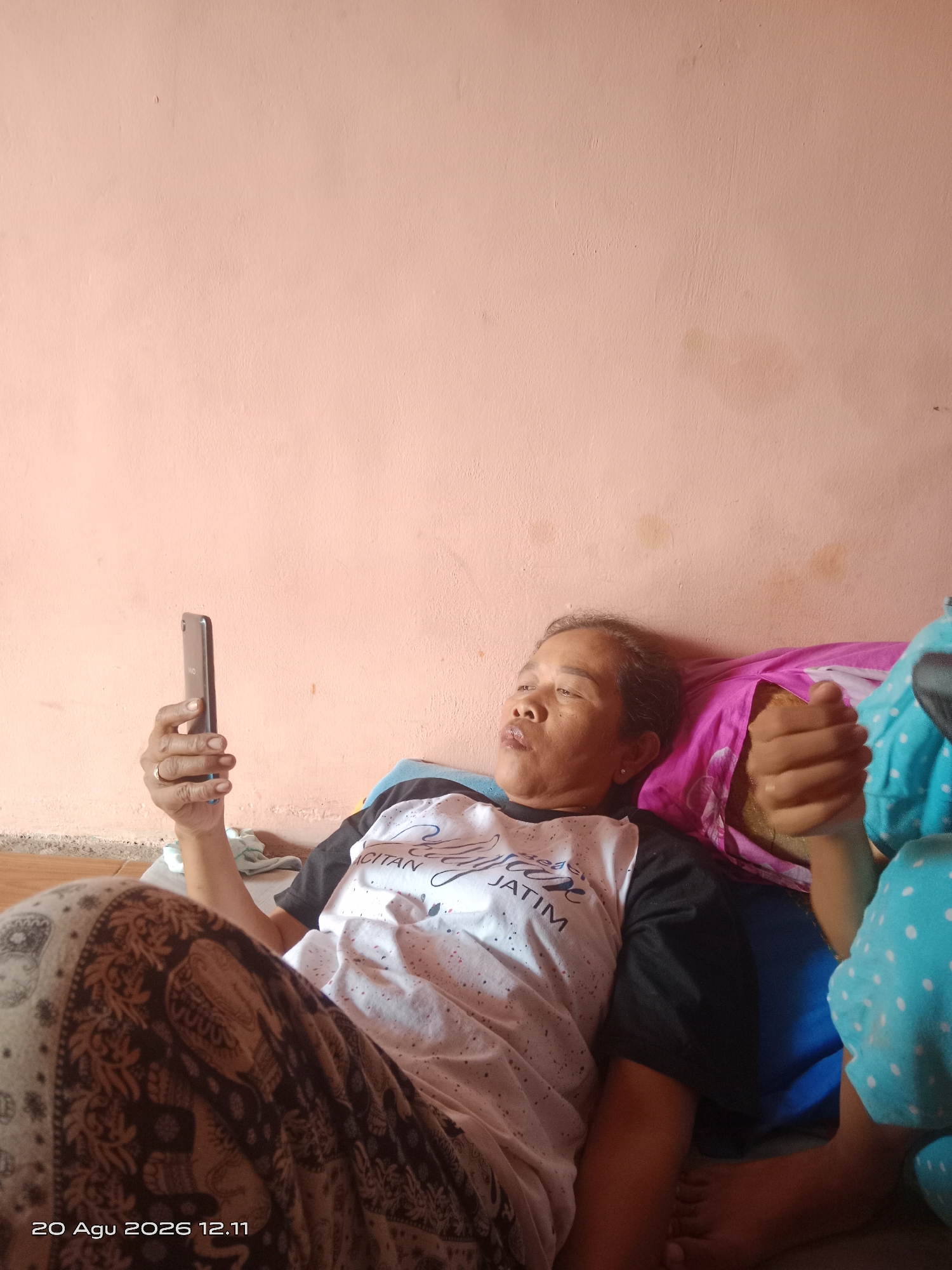 An indoor, medium shot captures a person lying on their side against a pale pink wall in what appears to be a residential setting in Kayen Kidul, Indonesia. The individual, with dark hair and fair skin, is wearing a white t-shirt with black sleeves and a patterned cloth covering their legs. They are holding a dark-colored smartphone in their right hand, angled towards their face as if viewing content. Their left arm is bent, resting near their head which is supported by a pink pillow. Another person, partially visible on the right side of the frame, is wearing a blue garment with white polka dots. The environment is sparsely furnished, with the focus on the individuals and their interaction with the phone. A timestamp in the bottom left corner reads "20 Agu 2026 12.11".