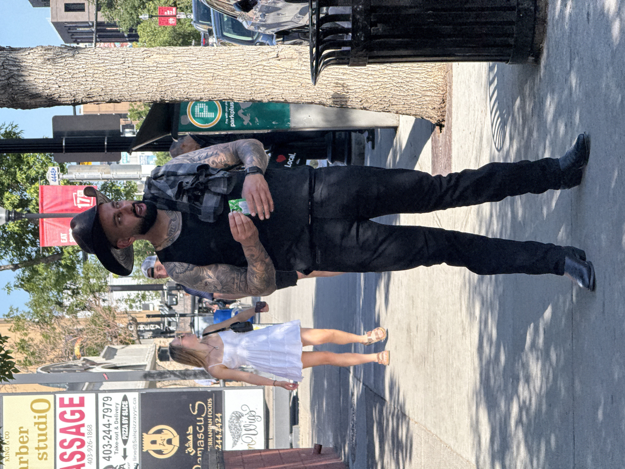 A man wearing a black cowboy hat, black sleeveless shirt, black pants, and black cowboy boots stands on a sidewalk in Calgary, Canada. He has tattoos on his arms and chest and is holding a small green packet in his right hand. To his left, a woman in a white sundress and sandals walks away from the camera, carrying a black purse. Behind the man, a tree trunk and a parking meter are visible. Signs for "King & Co. Barber Studio", "Massage", and "Damascus" are posted on a building to the left of the frame. The scene is lit by sunlight, casting shadows on the sidewalk.