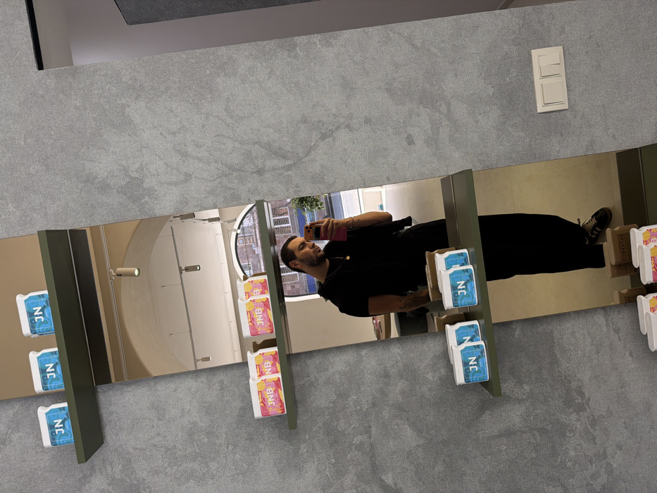 The image depicts a male subject taking a mirror selfie within a commercial interior in Yerevan, Armenia. The subject, reflected from the chest to the feet, is wearing a black short-sleeved top, black trousers, and brown low-top sneakers with white stripes. He holds a smartphone with a bright pink/orange case in his right hand, angled towards the mirror. A gold-colored necklace is visible.
The full-length mirror is mounted on a textured grey wall. Three dark green shelves are affixed to this wall, each extending into the mirror's reflection. The top and bottom shelves each display three white rectangular containers labeled "JN" with blue text on a blue background. The middle shelf holds three similar white containers, each labeled "JNB" with red text on a pink, yellow, and orange gradient background. To the right of the mirror, a white double light switch is mounted on the grey textured wall.
The mirror's reflection reveals a spacious interior with light-colored flooring and walls, illuminated by ceiling-mounted track lights. In the background of the reflection, an arched window or doorway provides an exterior view of an old, multi-story building constructed of brown-orange stone, featuring arched windows and balconies, consistent with local architecture.