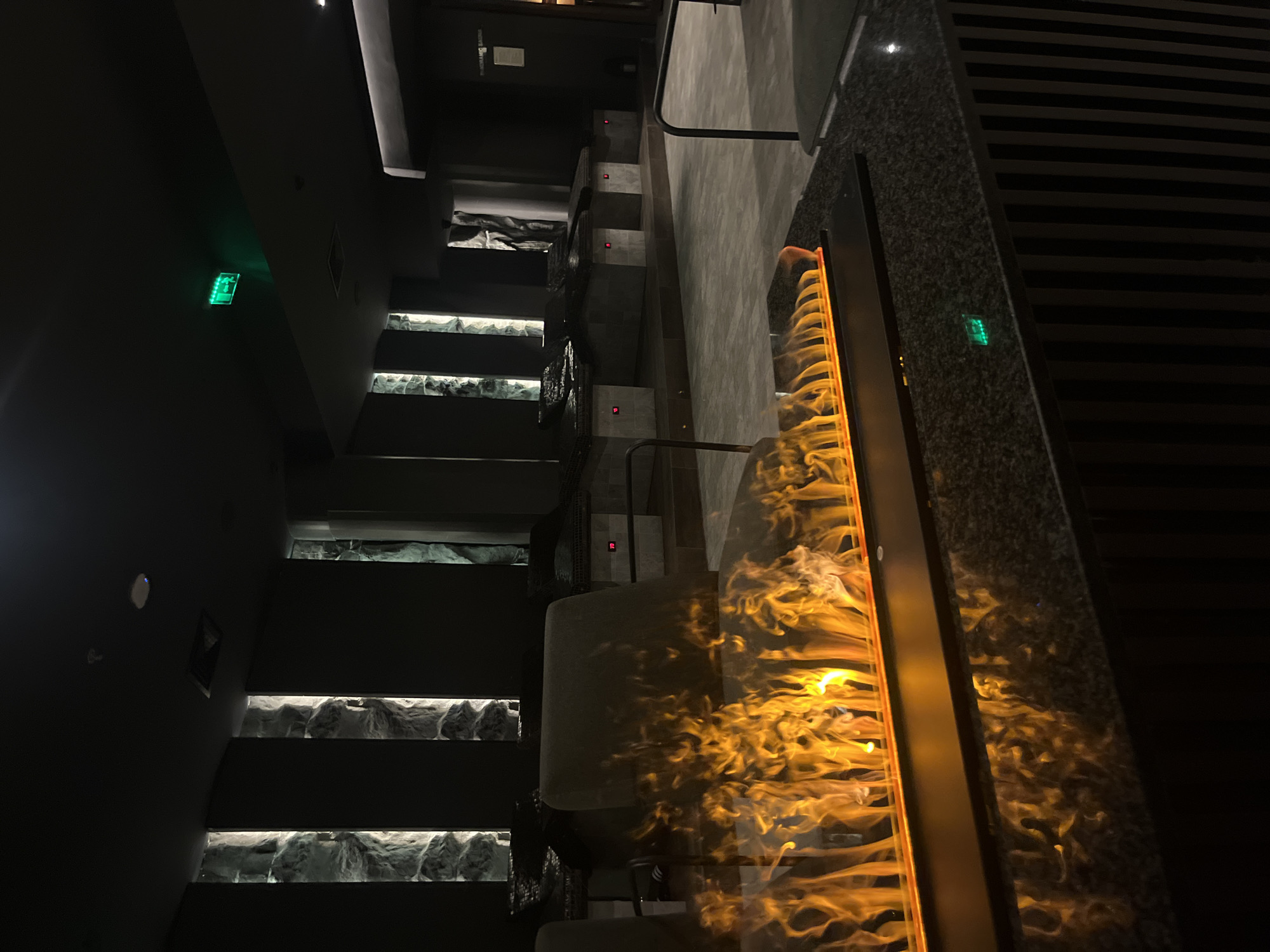 The image shows an interior space in Sofia, Bulgaria. A staircase with stone-look steps runs along the left side of the frame. Each step is illuminated from below. There are handrails on each side of the staircase. A vertical rectangular feature displaying an animated fire effect is on the right. To the left of the fire, there are upholstered seats. There is an "exit" sign in green on the wall.