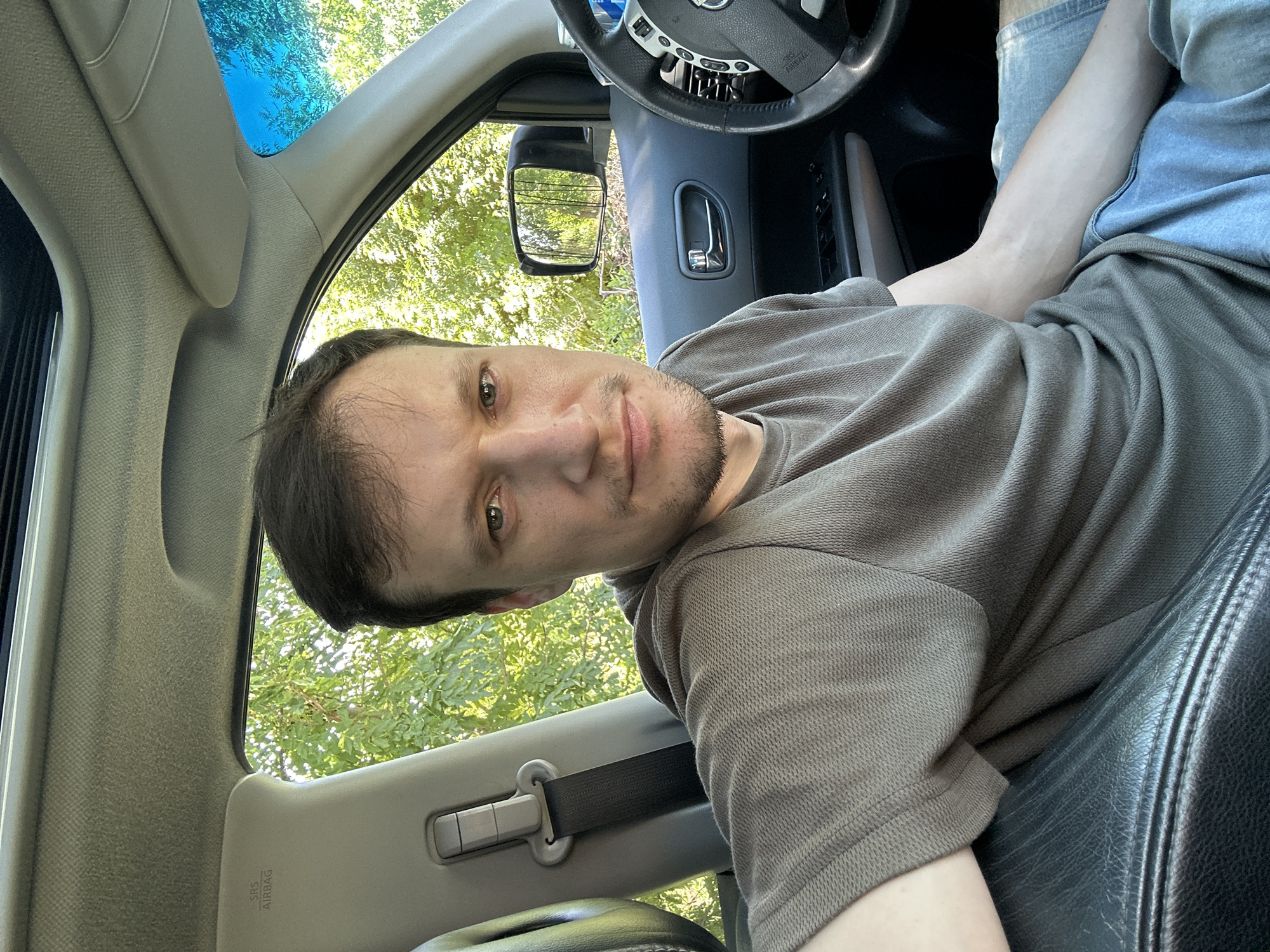 A man is sitting in the driver's seat of a car. The car's interior is visible, including a steering wheel, dashboard, and a side mirror. The man is wearing a grey t-shirt and light blue shorts. He is looking at the camera. Outside the car, trees and greenery are visible, suggesting a rural or suburban setting. The sky is blue and clear. The image appears to have been taken during daylight. The location is the city of Makiv, Ukraine.