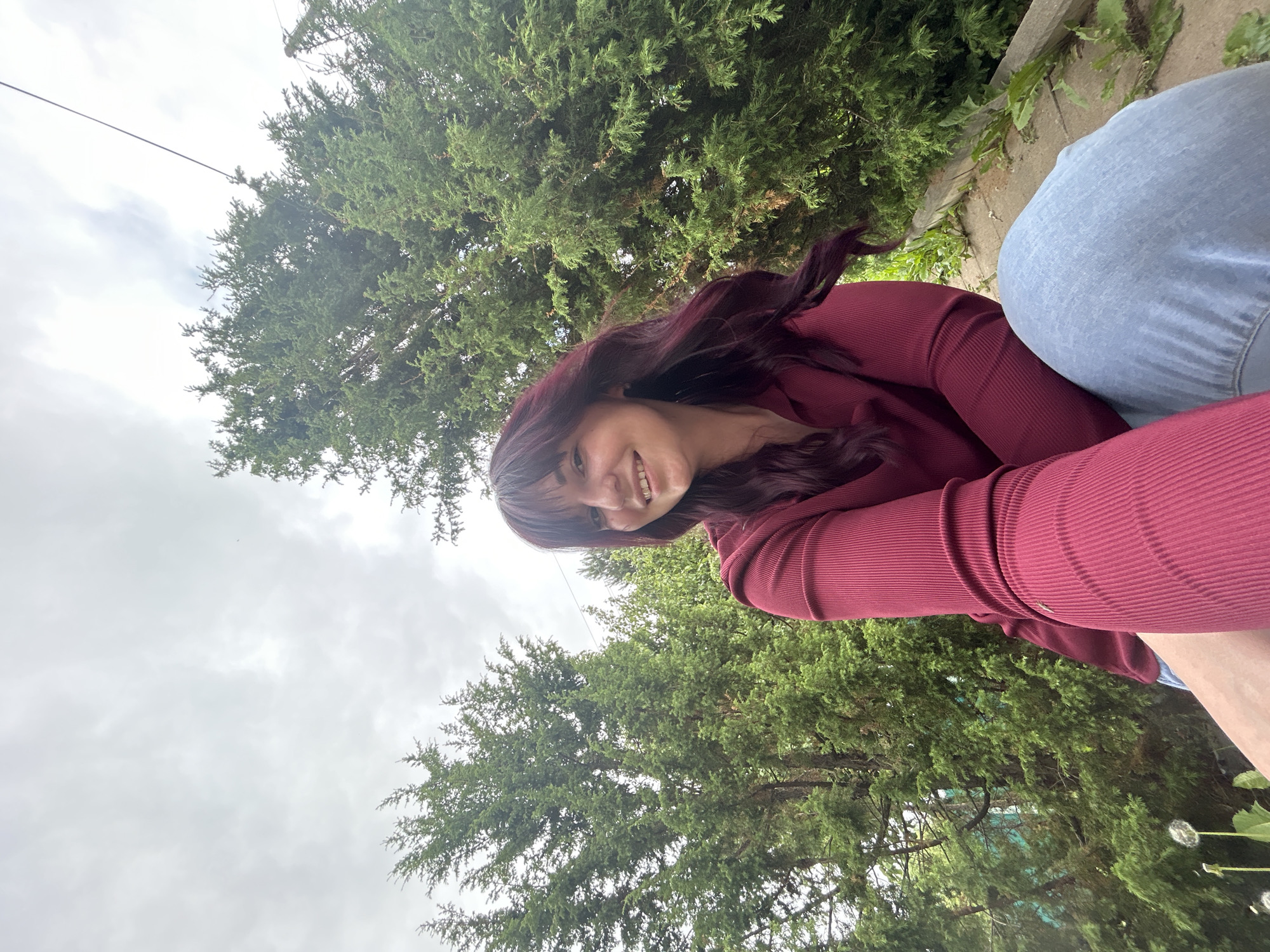 The image depicts a single female subject outdoors in the city of Фоки, Russia. The subject is a young adult woman with long, dark red hair and bangs, smiling directly at the camera. She is wearing a dark red, ribbed, long-sleeved top and light blue jeans. Her right arm is extended, indicating she is holding the camera for a selfie. She appears to be sitting or crouching on a raised surface, with her legs partially visible. The background consists predominantly of dense, tall green conifer trees under an overcast, light grey sky. A thin black power line or cable is visible horizontally across the upper right portion of the background, traversing the sky and trees. In the lower-left corner, some dandelion seed heads and green foliage are present. A concrete or stone edge is visible below the subject's right leg, with small plants growing along it.