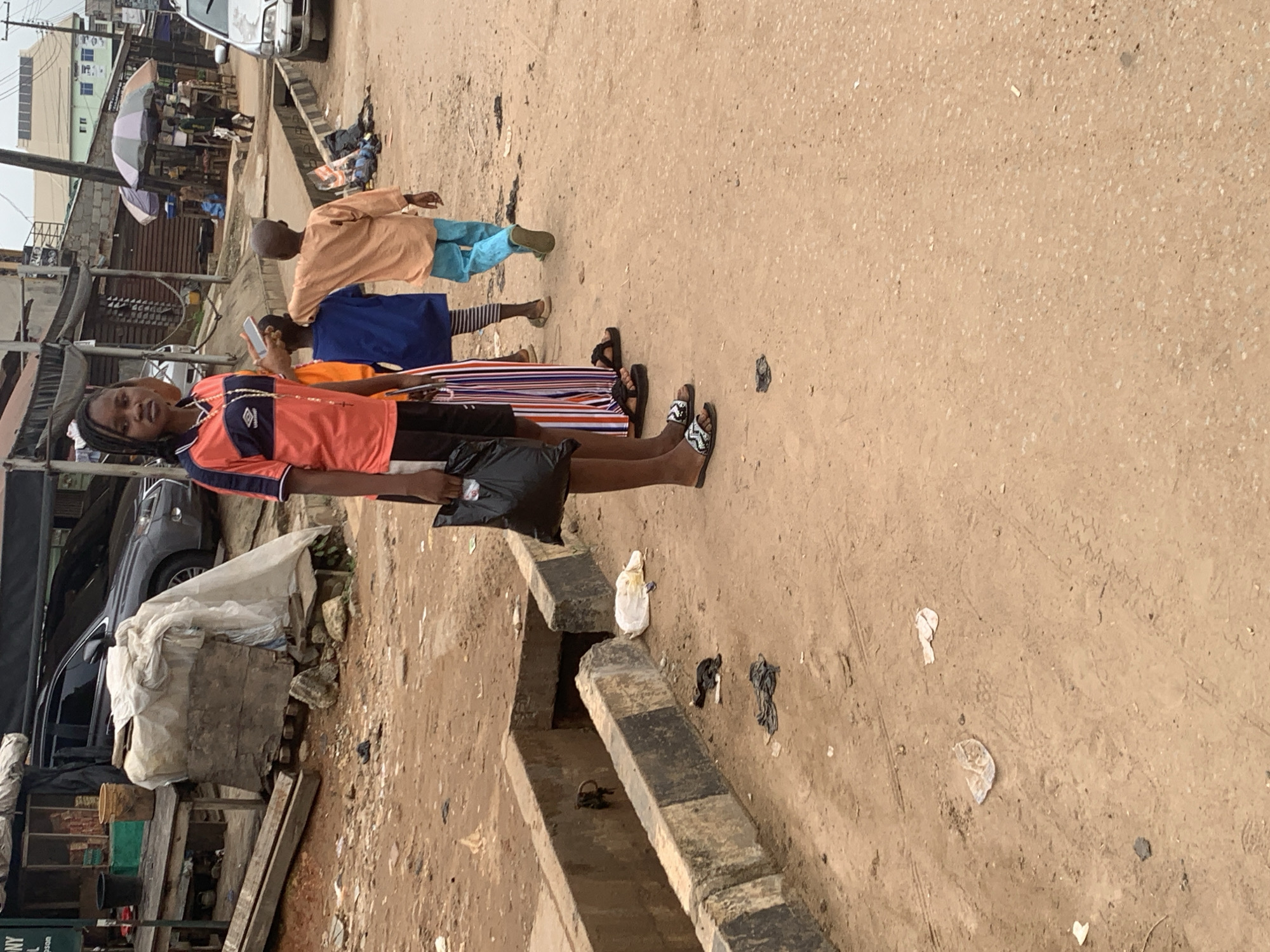 This image depicts a street scene in Shagamu, Nigeria, characterized by a dusty, unpaved road surface and a visible drainage channel. In the foreground, several children are present. Closest to the camera is a young girl wearing an orange and navy shirt and black shorts, holding a black plastic bag. To her right are two younger children; one in a blue shirt and striped leggings, and another in an orange shirt and light blue pants. The children appear to be standing on the side of the road.
The environment suggests a bustling, possibly commercial area, with market stalls and structures visible in the background. A dark-colored SUV is parked on the left side of the road, and a silver car is visible further down on the right. Scattered debris, including plastic bags and other litter, is present on the ground, particularly near the drainage channel, indicating a lack of consistent waste management. The overall impression is one of a typical urban street environment in Nigeria, with elements of daily life and commerce.
