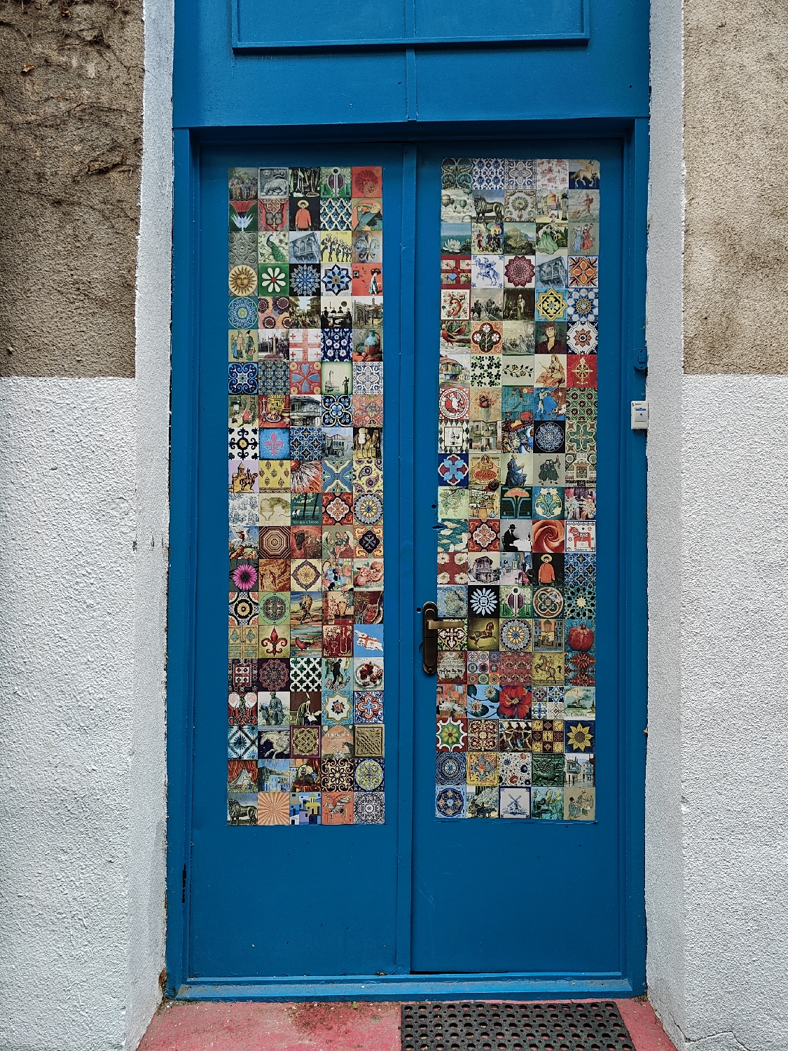 The image displays a vibrant blue double door, likely an entryway, set against textured walls. The left wall features a rough, off-white stucco with a brownish-grey patch at the top, while the right wall is solely off-white stucco. A red-painted concrete threshold and a dark, rubber doormat with circular perforations are visible at the base.
Both panels of the door are extensively covered with an eclectic collage of small, square decorative images, resembling ceramic tiles. These "tiles" display a wide array of designs and themes, including intricate geometric patterns like mandalas, stars, and cross-hatched motifs. Floral elements such as sunflowers, hibiscus, and various leafy patterns are interspersed with diverse animal figures like peacocks, horses, and birds. Many tiles depict human figures engaged in various activities, religious iconography, historical scenes, and mythological creatures, often rendered in folk art or medieval styles. Architectural elements, heraldic symbols like the fleur-de-lis, and abstract patterns contribute to the rich visual tapestry.
A single, ornate dark-colored door handle is centrally located on the right door panel. Above the double doors, a solid blue panel completes the frame. The scene is illuminated by soft, natural light, suggesting daytime without harsh shadows. This artistically adorned entrance is situated in Tbilisi, Georgia. No clear, legible text is visible within the intricate designs. The overall impression is one of artistic expression and individuality.
