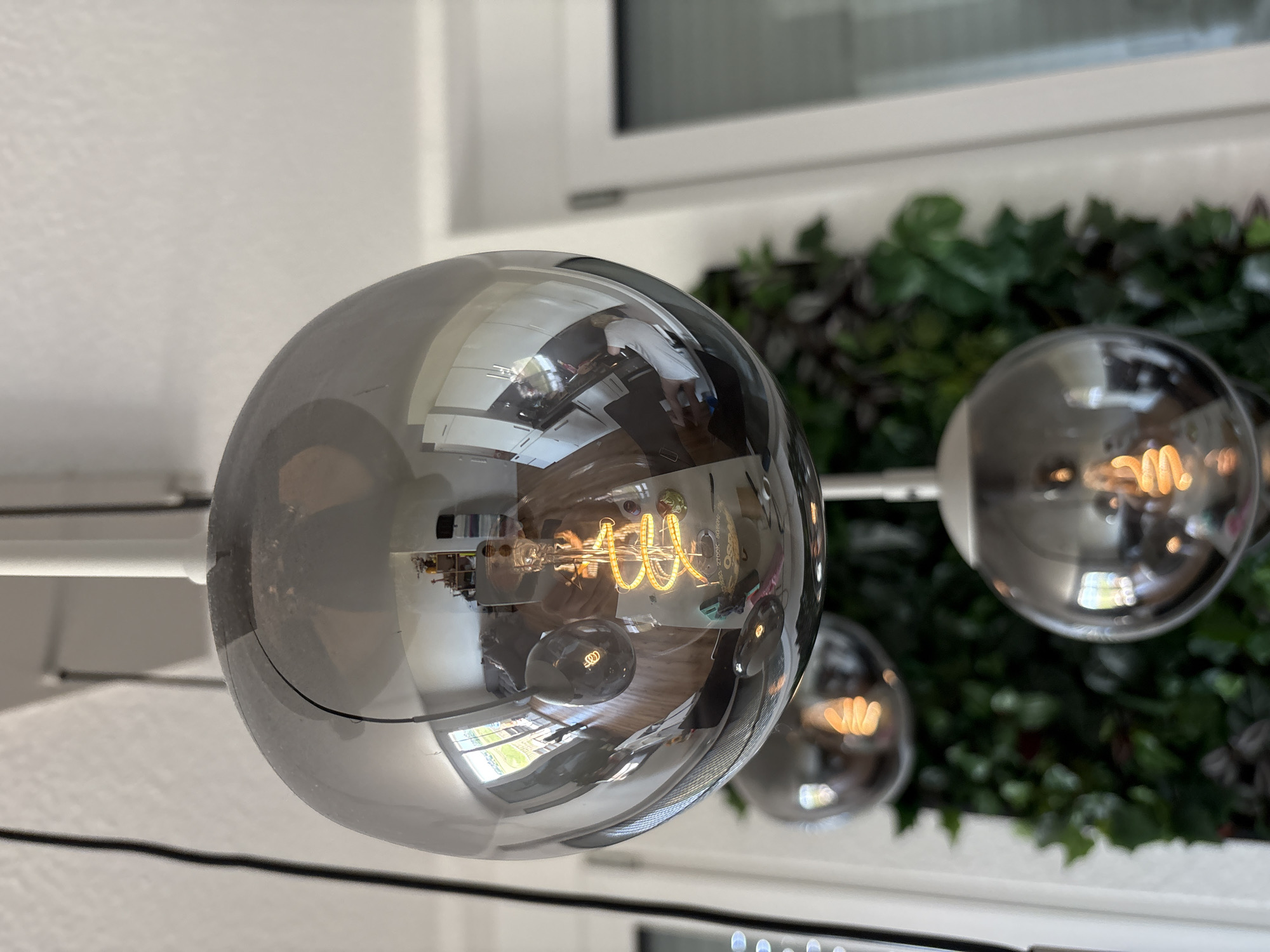 A close-up shot captures a cluster of three spherical, mirrored pendant lights hanging from white rods against a white ceiling. The primary light, in the foreground, is a large, dark gray sphere with a highly reflective surface. Inside this sphere, a glowing, spiral filament lightbulb emits a warm, yellow light. The reflection on the sphere's surface reveals an interior scene with wooden floors, white cabinets, and what appears to be a kitchen area. A person is visible in the reflection, wearing a light-colored shirt and holding a mobile phone. Two other smaller, mirrored pendant lights are visible below and slightly to the side of the main light, also containing glowing filament bulbs. In the background, a vertical green wall or plant display is partially visible, along with a white door frame and a window. The scene is indoors, with a bright, natural light source coming from outside. The location is Bönnigheim, Germany.