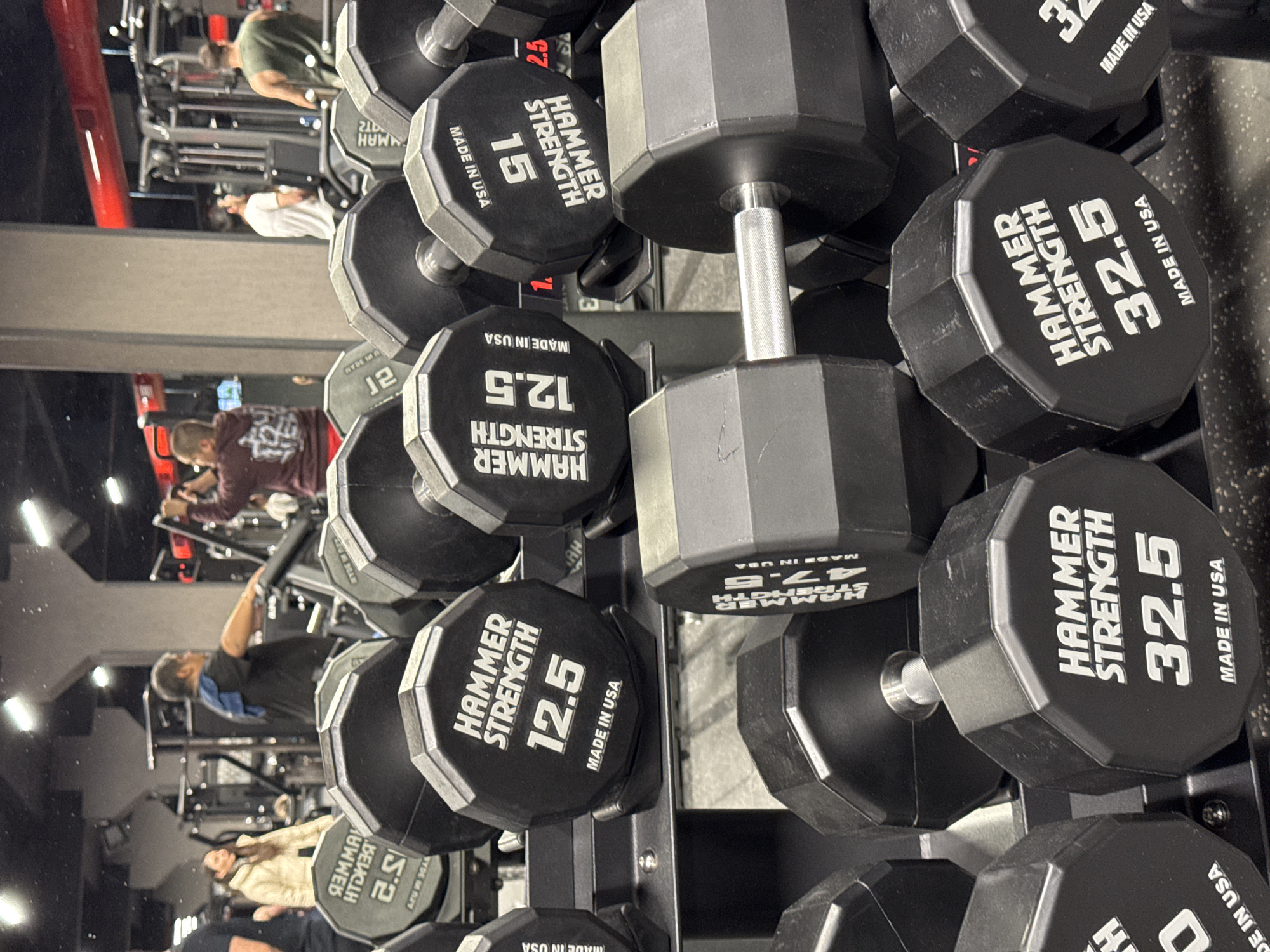 The image shows a rack of Hammer Strength dumbbells in a gym. The dumbbells range in weight from 12.5 to 47.5. Several people are visible in the background, engaged in various workout activities. The gym has weightlifting equipment and workout machines. The location is not specified as Yerevan, Armenia, in the image.