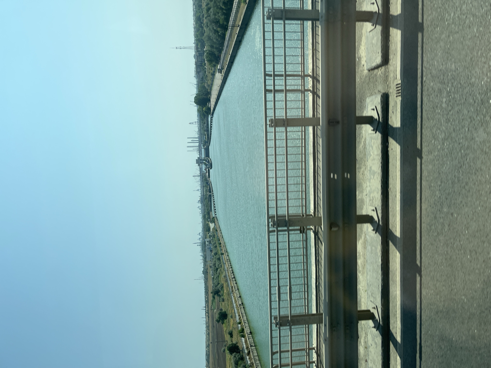 The image shows a view from a bridge overlooking a wide, calm canal. The canal is lined with concrete walls and on the far side, there is vegetation and industrial structures, including cranes and towers, suggesting a port or industrial area. The sky is clear and blue. In the foreground, a metal railing and the side of the bridge are visible. The scene is likely located in Agigea, Romania, as it depicts the Agigea Canal, a vital waterway connecting the Black Sea to the Danube-Black Sea Canal.