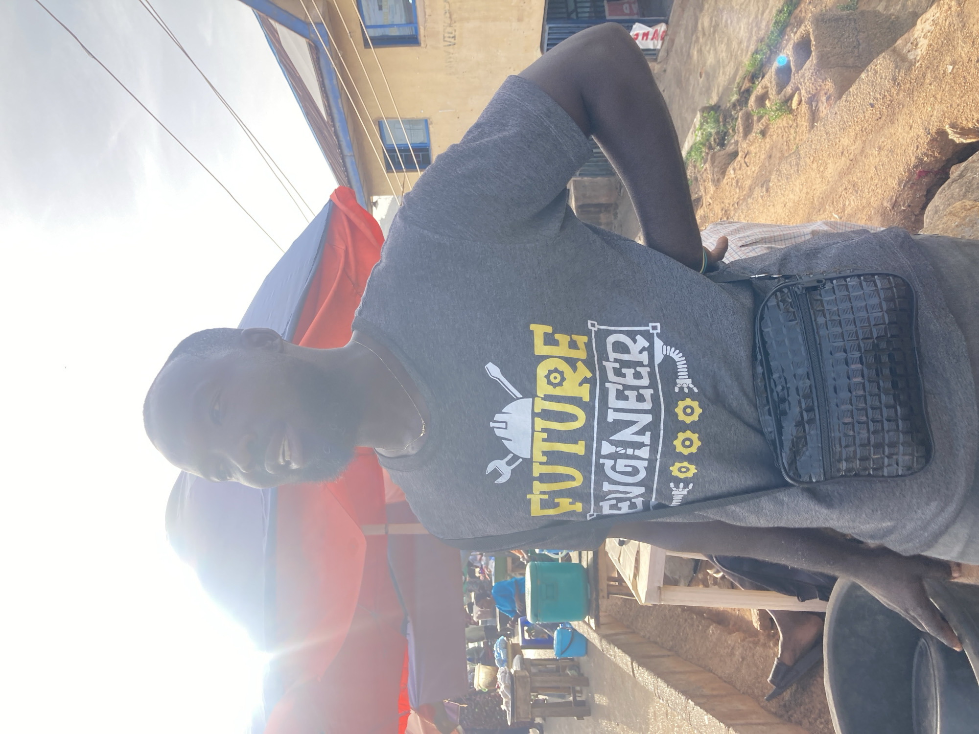 A man wearing a "Future Engineer" t-shirt and a black bag is standing outdoors in Gwong, Nigeria. The background shows buildings and street activity. The lighting is bright, possibly midday sun.