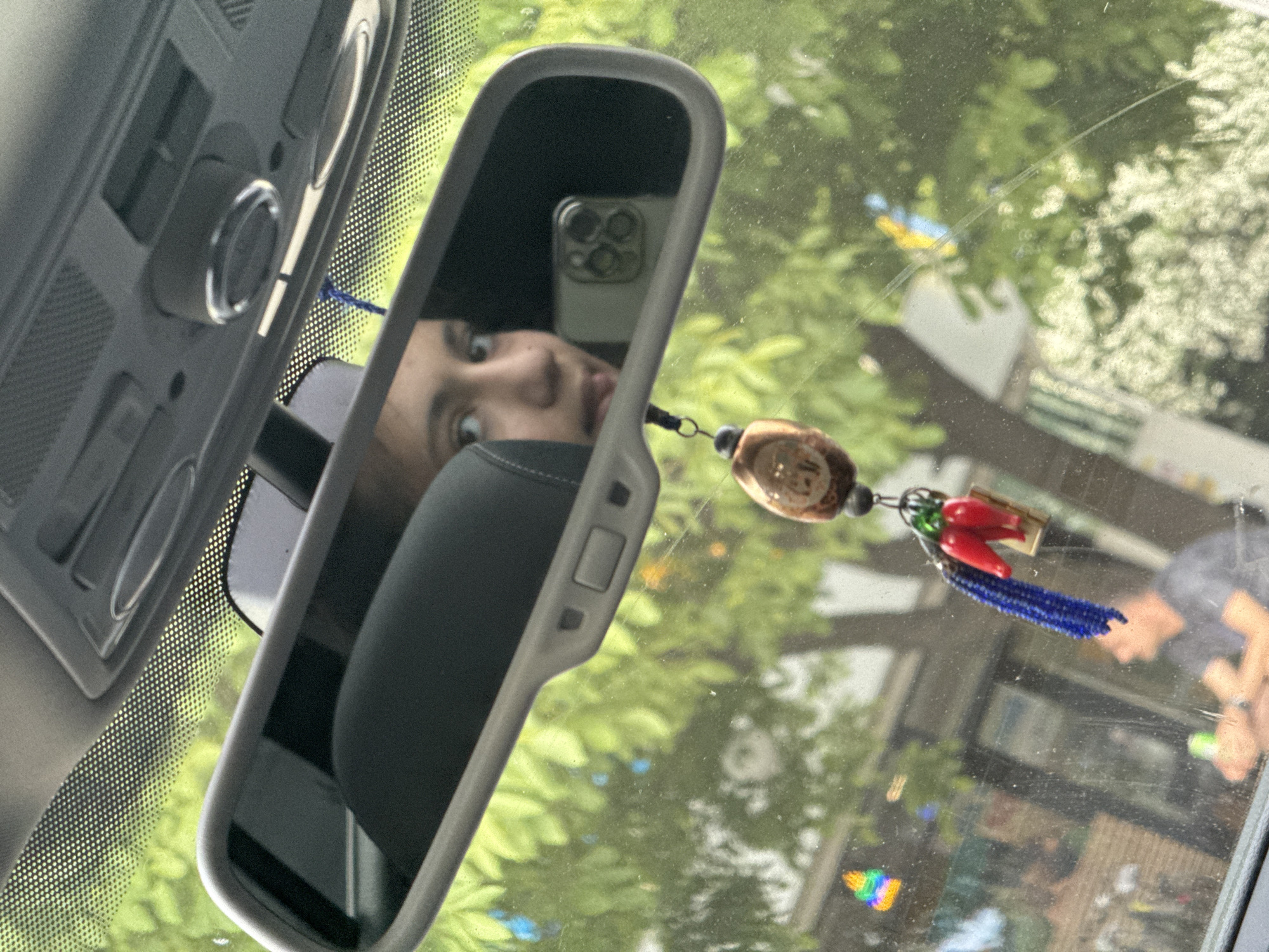 The image captures a view from within a vehicle's interior, looking forward through the windshield, situated in Suprunivka, Ukraine. The primary subject is the rearview mirror, which reflects a young person's partial face, including their eyes, nose, and mouth, positioned towards the left side of the mirror. To the right within the reflection, a light-colored smartphone, identified by its distinct triple-camera array, is clearly visible, indicating the individual is taking a selfie.
A decorative charm hangs from the rearview mirror, composed of a spherical, dark brown element at the top, three red chili pepper-shaped pendants, and a tassel of small blue beads.
Portions of the car's gray overhead console and headliner are visible above the rearview mirror, along with the dotted fritted band on the windshield.
Through the windshield, a daylight exterior scene is visible, characterized by dense green tree foliage. A blurry individual, appearing male, dressed in a gray shirt, is observed in profile in the lower portion of the external view, seemingly holding an item. The windshield surface displays minor dust and reflections, including a small rainbow-colored object at the bottom-left and a blue and yellow item further up.