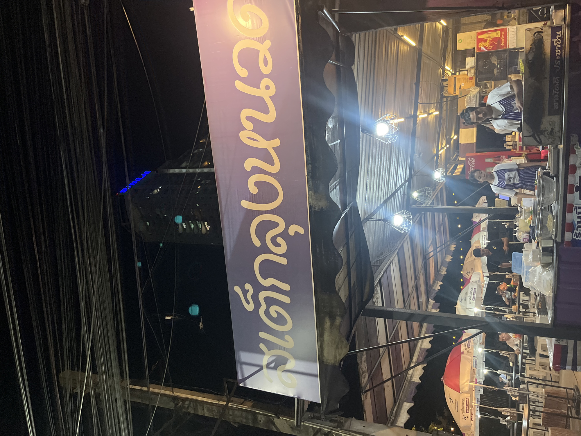 The image captures a bustling night scene in Pattaya City, Thailand. A purple banner with golden Thai script prominently displays "สเต็กลุงหนวด" (Steak Lung Nuat) and "เด็กลุงหนวด" (Lung Nuat's Child), likely indicating a local restaurant. The scene is illuminated by multiple hanging lights, casting a bright glow on the surroundings.
The backdrop features corrugated metal structures, suggesting a semi-permanent setup. People are visible, including individuals wearing striped aprons, presumably staff members preparing food behind a counter. Underneath the roof are people dining and enjoying the restaurant. A white and red umbrella is seen in the foreground.
In the background, a tall building with blue lights can be distinguished. Overhead, a web of electrical wires adds to the urban landscape. Other food branding/advertising includes Coca-Cola and Samaddee Prachacheun on the awning. The image captures a vibrant street food atmosphere at night.