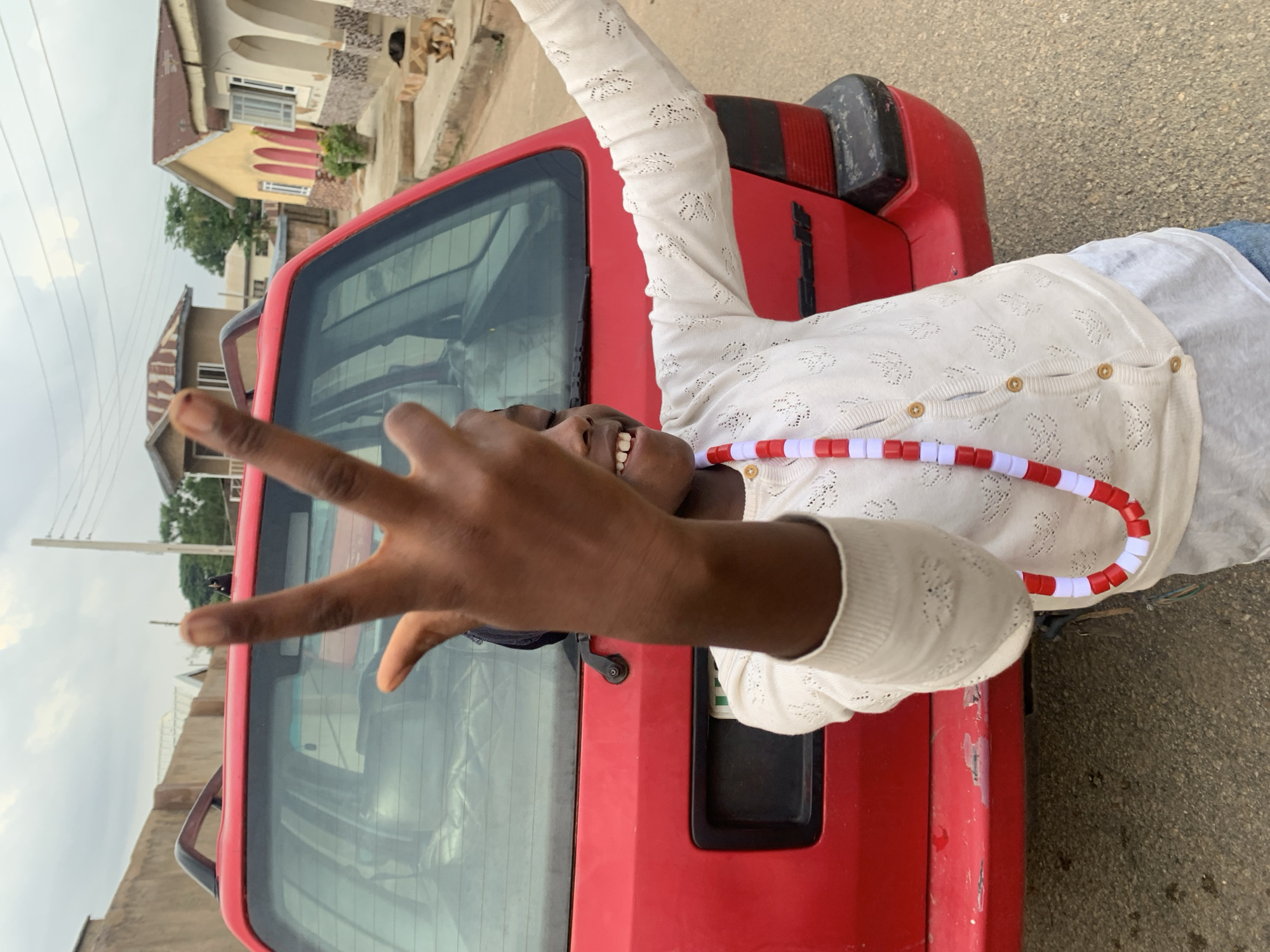 The image shows a young Black person lying down on top of a red car. The person is wearing a white knitted cardigan and blue jeans. They are smiling and holding up their left hand in a peace sign. A necklace of red and white beads is visible around their neck. The car appears to be parked on a street in a residential area, with houses visible in the background. The sky is overcast, suggesting it might be a cloudy day. The overall mood of the image is cheerful and playful. There is no visible text in the image. The context provided indicates the location is Jos, Nigeria.