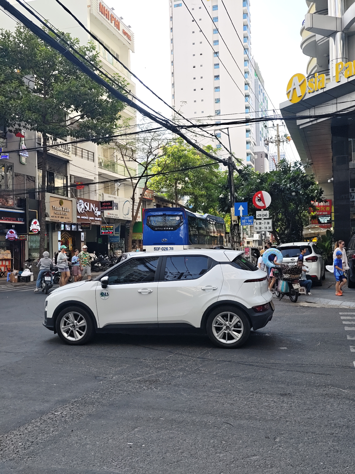 The image captures a street scene in Nha Trang Ward, Vietnam, characterized by its bustling urban environment. A white car, possibly a taxi, is prominently positioned in the foreground, angled slightly towards the viewer and facing left. The car appears to be stopped or moving slowly on a paved road, which shows signs of wear and tear.
Behind the car, a blue bus with the branding "VIETNAM UNIVERSE" is visible, suggesting public transportation is operating in the area. The bus also displays a license plate. To the right of the bus, traffic signs are present, including a directional arrow and a sign with a red circle and a diagonal line, indicating a restriction or prohibition. Further to the right, people are seen walking on the sidewalk, with one individual in the foreground wearing a blue shirt and shorts, and another, a woman with blonde hair, in the background.
The background is dominated by multi-story buildings, some of which are modern high-rises with numerous windows, while others appear to be older structures with balconies. One building displays a large yellow "A" logo, possibly indicating a business or establishment. A prominent sign for "NHI PHU HOTEL" is visible on a white building in the upper left portion of the image. Numerous electrical wires are strung across the scene, a common sight in many urban areas. Trees with green foliage are interspersed between the buildings, adding a touch of nature to the urban landscape. Several motorbikes are parked along the side of the street, indicating a popular mode of transport. The overall impression is one of a lively street with ongoing activity.