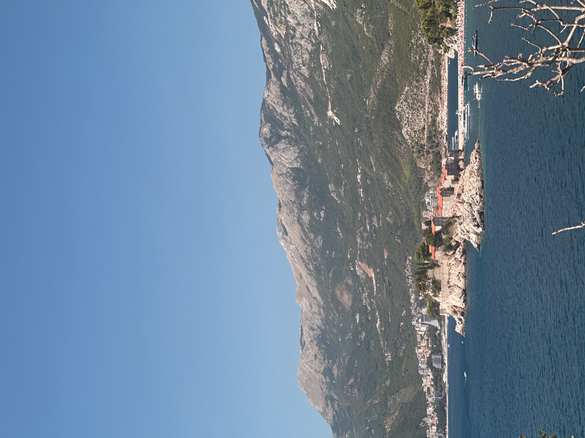 The image shows a clear blue sky above a mountainous landscape and a coastal town. In the foreground, a body of deep blue water occupies the lower portion of the frame. A rocky islet, identified as Sveti Stefan in Montenegro, is situated in the middle ground. This islet is dotted with buildings featuring red tile roofs, suggesting it is a historical or resort area. A narrow land bridge connects the islet to the mainland.
Along the coastline to the left of the islet, a town with numerous buildings, including hotels and residential structures, is visible. The mountainous terrain behind the town is covered in green vegetation and appears rocky in places. Several boats are present in the water, including yachts and smaller vessels. A beach area with numerous people can be seen further along the coast to the right of the islet. In the extreme foreground, the skeletal branches of a bare tree are visible.