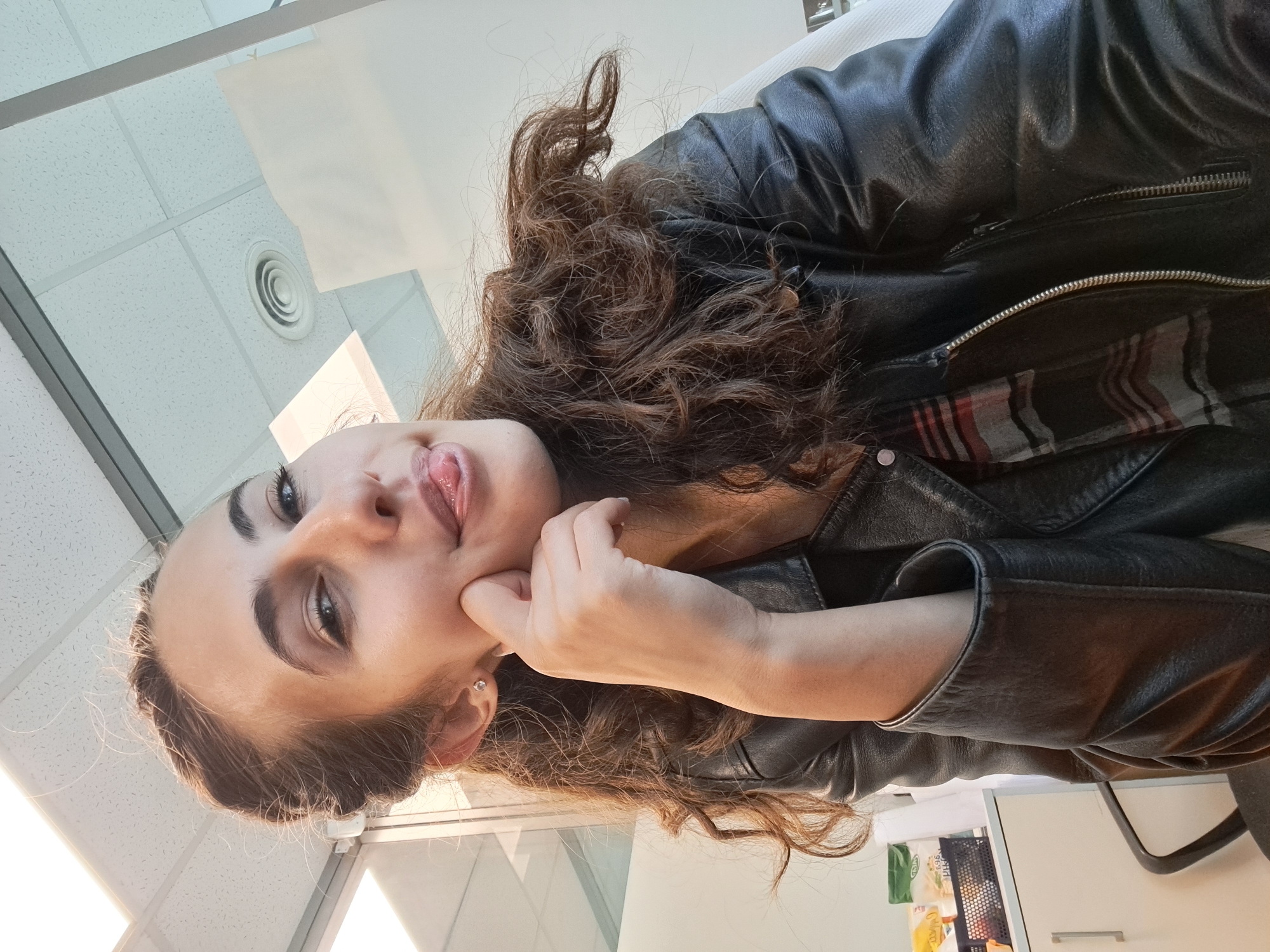 A young woman is pictured in a selfie. She is indoors, likely in an office environment, with a white ceiling featuring fluorescent lighting and a circular air vent visible. She has long, curly brown hair and is wearing a black leather jacket over a plaid shirt. Her right hand is touching her cheek, and her tongue is sticking out playfully. The background includes a desk with some items on it, a filing cabinet, and a glass partition. The overall lighting is bright and appears to be from overhead sources.