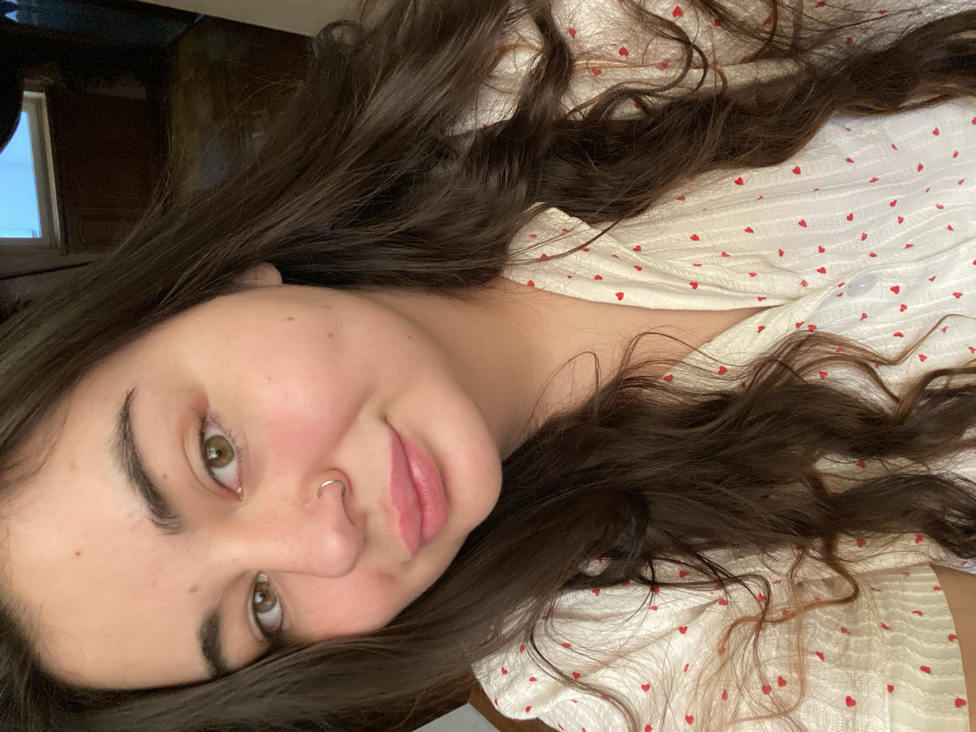 A close-up, tilted shot features a person with fair skin, dark brown hair, and green eyes. A silver hoop is visible in their left nostril. Their hair, styled in loose waves, is partially spread across a white fabric with small red heart patterns. The person is smiling subtly. The background is out of focus, showing a dark object and a window with daylight visible through it. The location is unspecified.