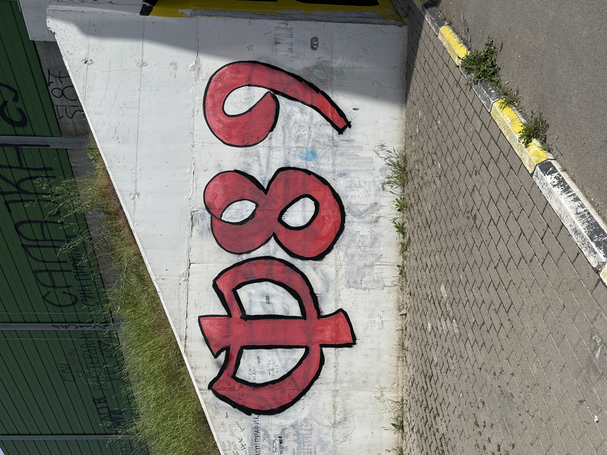 A close-up, eye-level shot displays a section of a concrete structure in Novi Sad, Serbia, adorned with vibrant red graffiti numbers arranged vertically: '6,' '8,' and a symbol resembling an uppercase 'D' with a horizontal line through its center. These numbers are outlined in black, adding definition to their bold presence against the concrete background. The concrete structure appears to be a retaining wall or part of a bridge support, with various other, smaller graffiti markings visible on its surface. To the left of the concrete structure is a green corrugated metal fence, and below the fence, grass grows along the base of the structure. To the right of the concrete structure is a brick walkway, with a yellow and black painted curb separating it from a gray paved road. The scene is bathed in sunlight, creating shadows and highlights across the surfaces, adding depth to the composition.