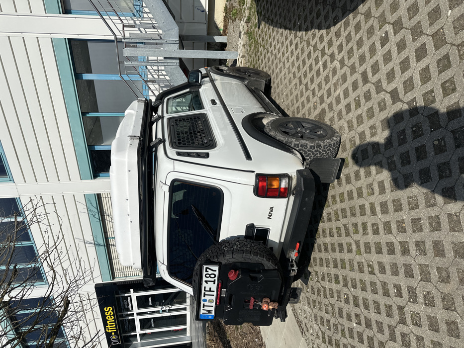 A white Lada Niva 3-door SUV, heavily modified for off-road and overland travel, is parked on an interlocking concrete paver surface in front of the "pro-fitness" gym located at Schaffhauser Str. 3, 79798 Jestetten, Germany.
The vehicle is positioned with its rear and right side facing the camera under bright sunlight, casting prominent shadows, including the photographer's shadow in the foreground. It features a white body, black fender flares, black side steps, and black multi-spoke wheels fitted with aggressive off-road tires. A large white hard-shell rooftop tent is mounted on a black roof rack.
The rear of the vehicle displays a spare tire mounted on a carrier to the left, with a black jerry can attached to its left side, from which a small plush monkey toy dangles. A German license plate, "WT TF 187", is centrally mounted above the spare tire. The robust black rear bumper includes a red tow hook shackle and a red-tipped locking mechanism. Black mud flaps are present behind the rear wheels. The rear side windows are covered with black metal grids.
In the background, the two-story "pro-fitness" building, with light-colored horizontal siding and prominent blue window frames and trim, is visible. An external metal staircase with a grey railing ascends along the building's right side. Bare tree branches are visible in the upper left portion of the frame.