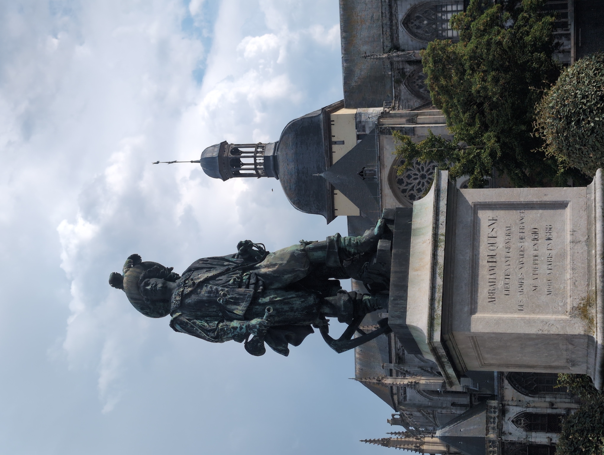 The image depicts a weathered bronze statue of Abraham Duquesne, a prominent figure in French naval history, standing on a stone pedestal. Duquesne is depicted in military attire, complete with a feathered hat and holding a baton. The statue is located in front of the Church of Saint-Jacques in Dieppe, France, evident from the architectural style of the building in the background, which includes Gothic spires and a large rose window. The sky above is cloudy, with a mix of sunlight and shadows casting across the scene. The base of the statue bears an inscription detailing Duquesne's life, stating he was born in Dieppe in 1610 and died in Paris in 1688. Lush green foliage surrounds the base of the monument and the church.