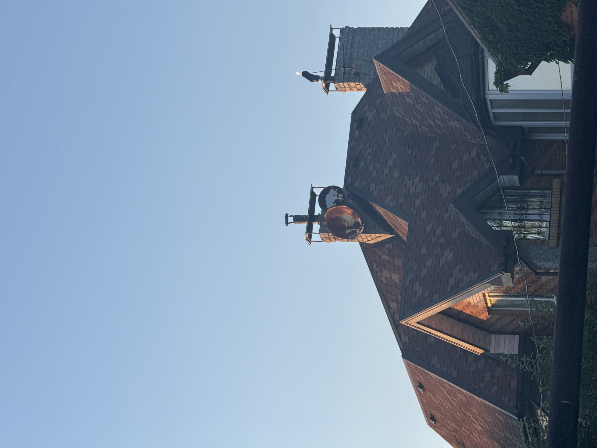 In Obukhivka, Ukraine, the image captures the upper section of a residential building with a prominent brown asphalt shingle roof. The roof features multiple gables, some with decorative triangular overhangs, and a section covered in ivy. Two chimneys are visible on the rooftop. The left chimney appears to be a brick structure topped with a metal cap and an aged, rusty satellite dish. The right chimney is also brick, with a metal cap, and supports a dark cylindrical object with a white tip, possibly a light fixture. The sky above is clear and a pale blue. In the foreground, a dark horizontal element, possibly a fence or railing, partially obscures the view of greenery. The lighting suggests it is either early morning or late afternoon, with warm sunlight illuminating the roof surfaces.